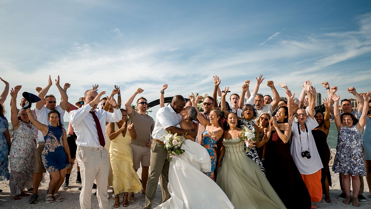 Joyful beach wedding celebration with bride, groom, and guests cheering with raised arms under a blue sky.