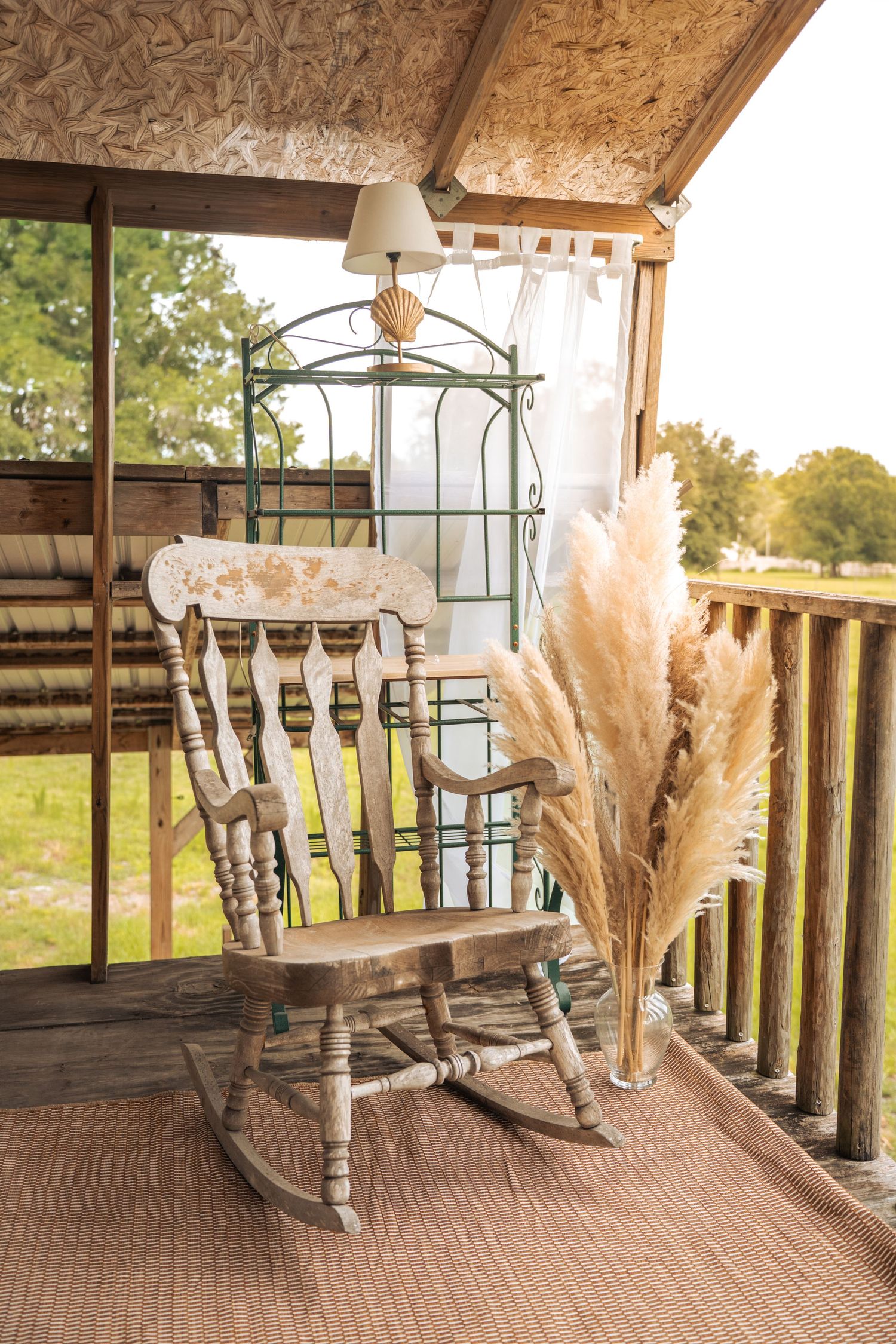 Rustic wooden rocking chair on a country porch with pampas grass, sheer curtains, and a vintage lamp in warm sunlight at Farmettes Photo Stu