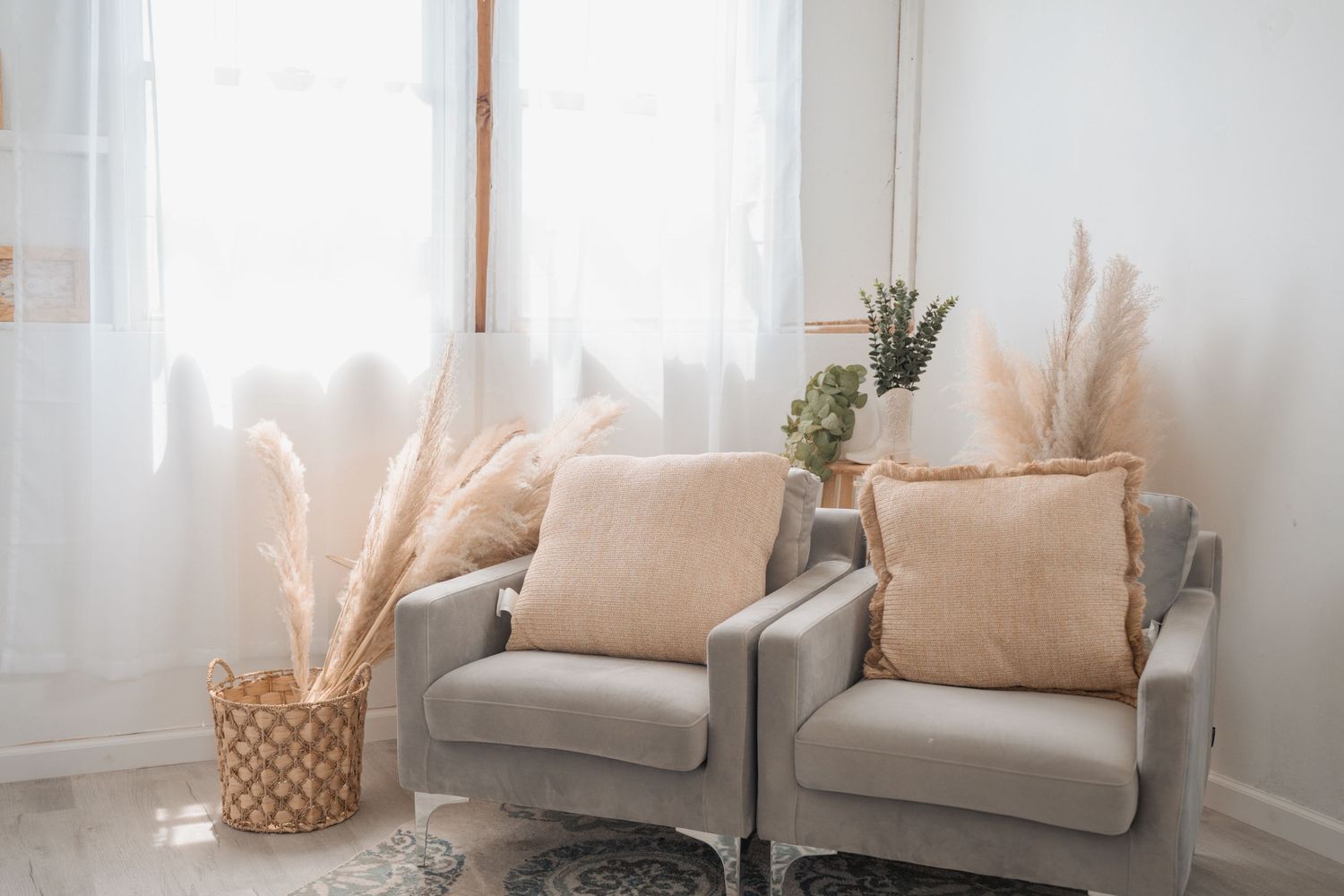 Two gray accent chairs with beige pillows beside pampas grass decor near a bright window in a modern living room.