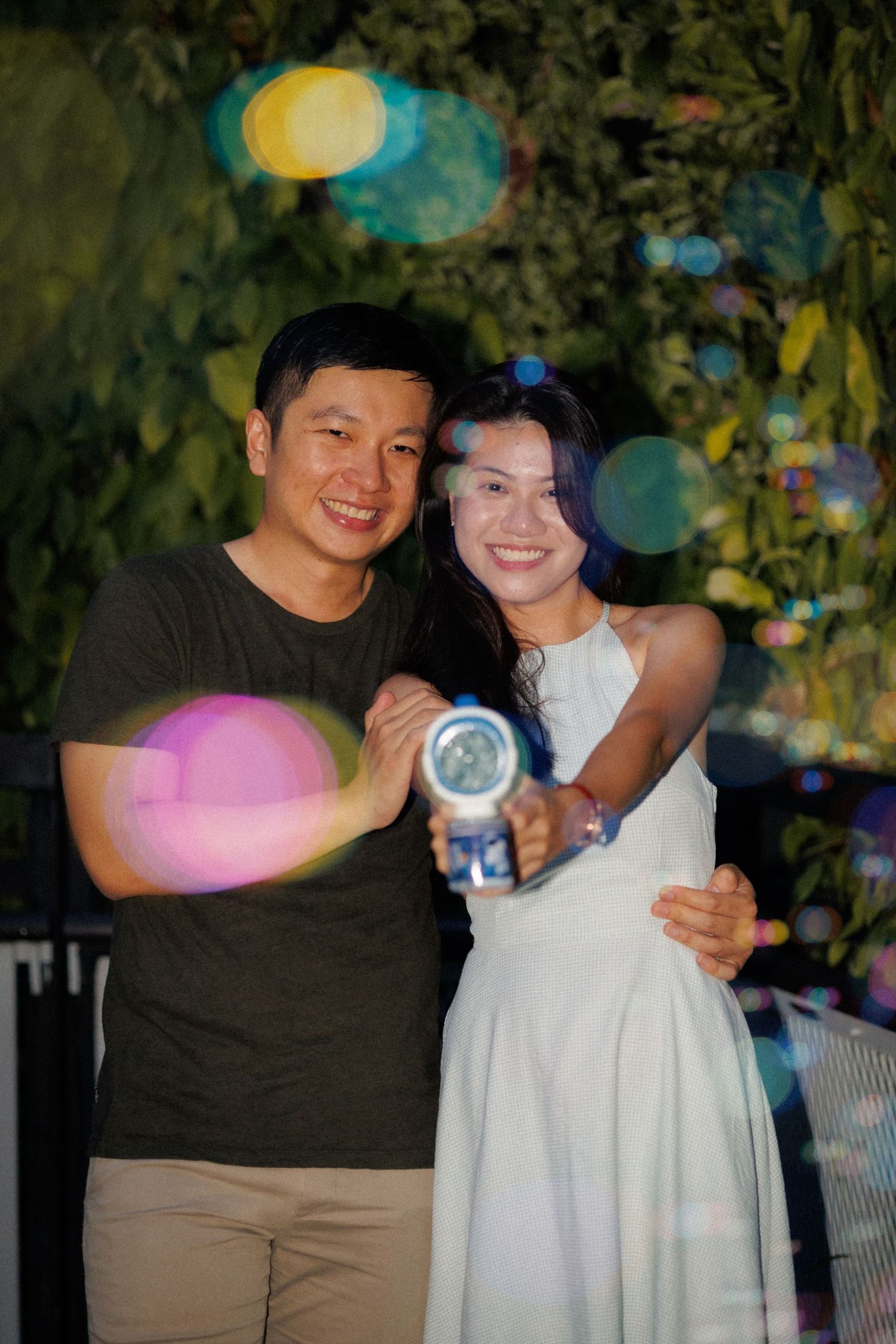 Couple in casual attire posing together at night with decorative lights in background.