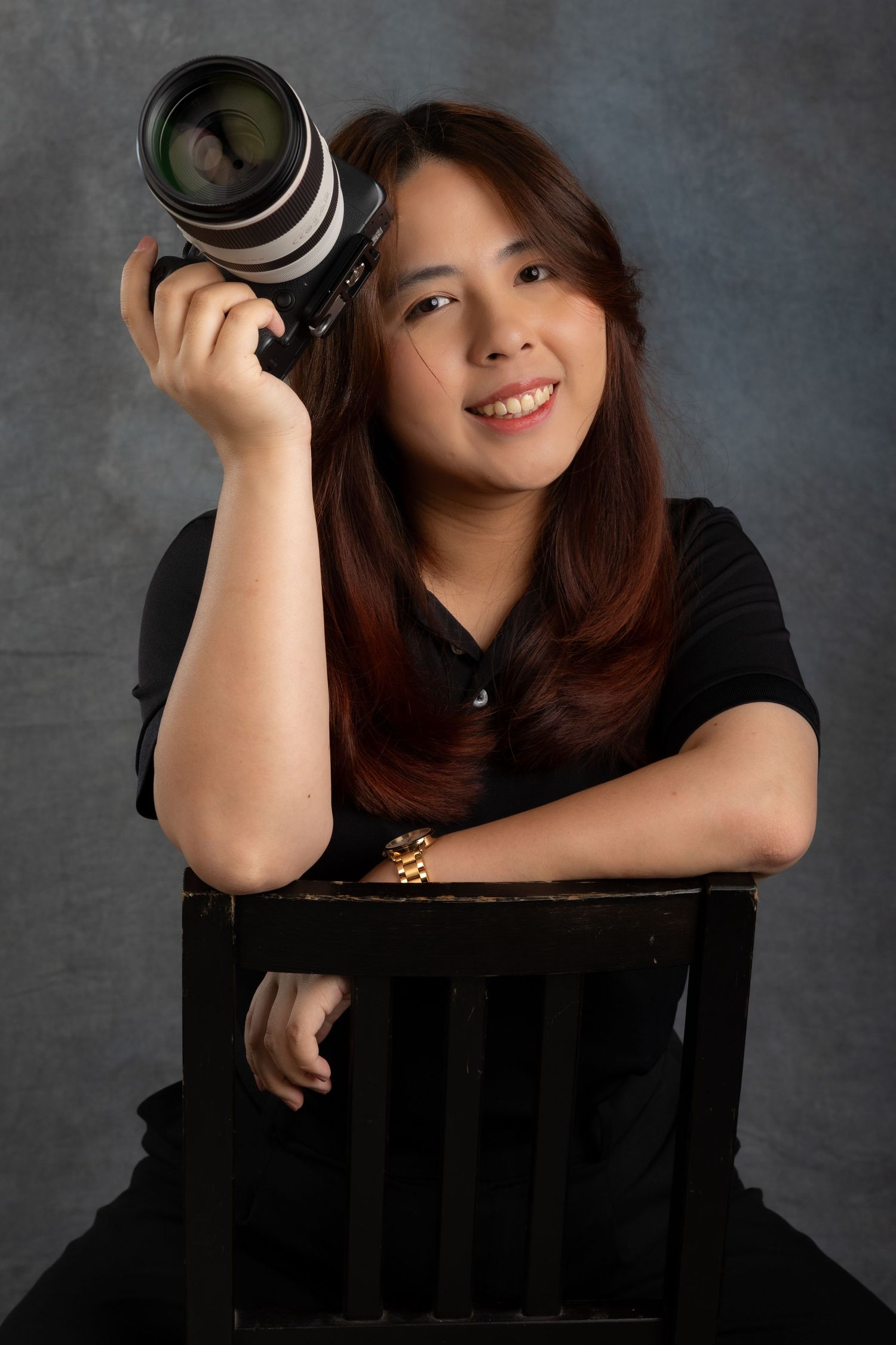 Photographer in black shirt poses with professional camera while leaning on wooden crate against gray backdrop.