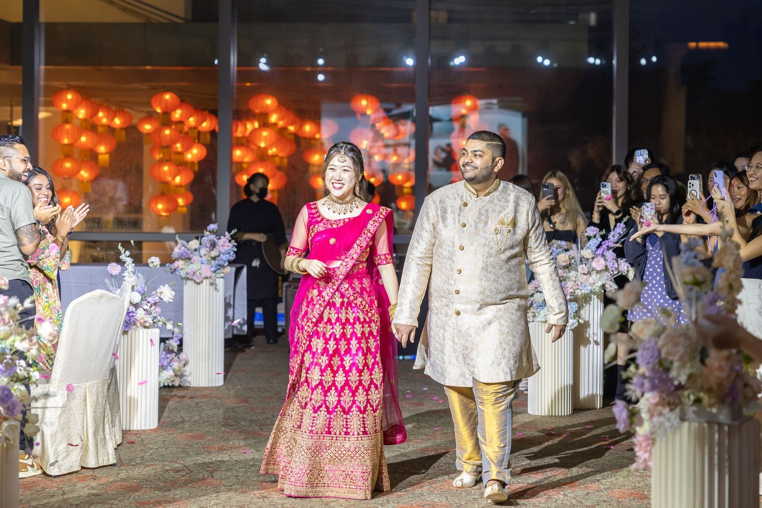 Couple in traditional South Asian wedding attire walking down decorated aisle.