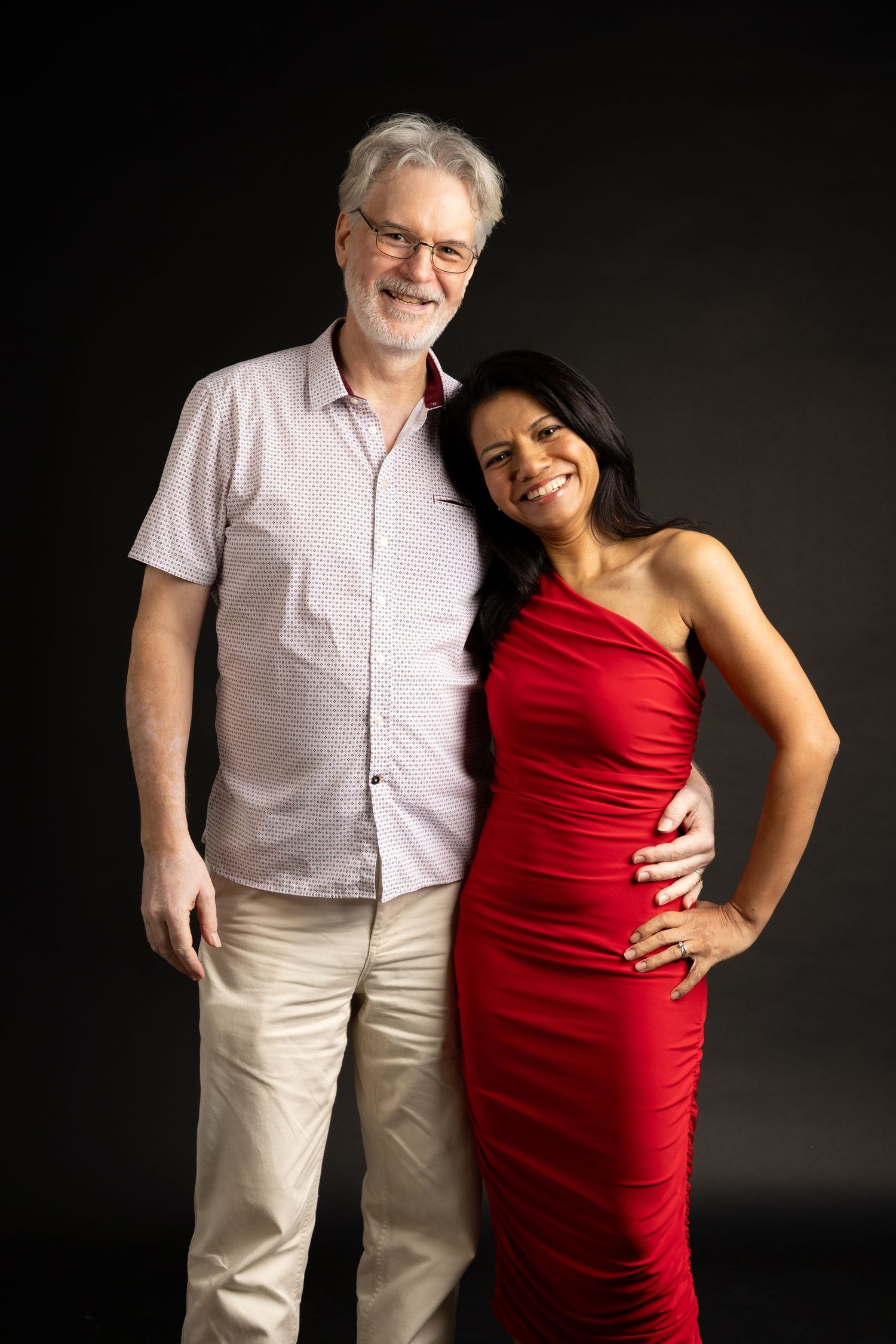 A couple poses together against a dark background, with her in red and him in white.