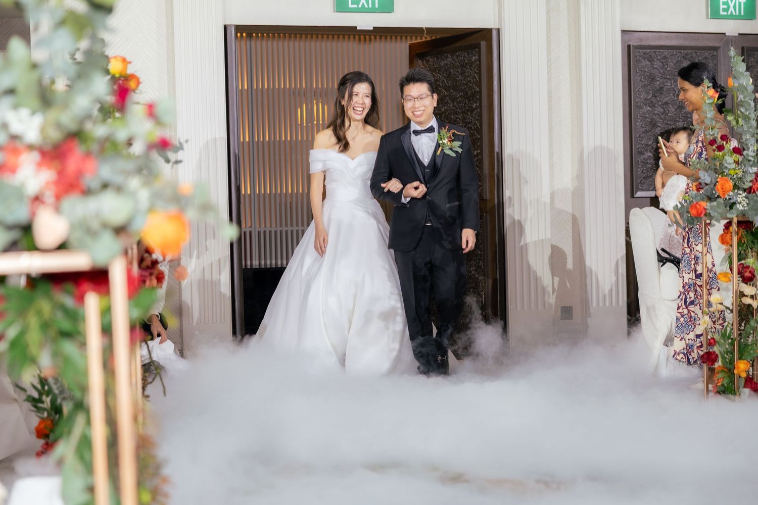 A couple in wedding attire poses in a wooden doorway decorated with flowers and eucalyptus.