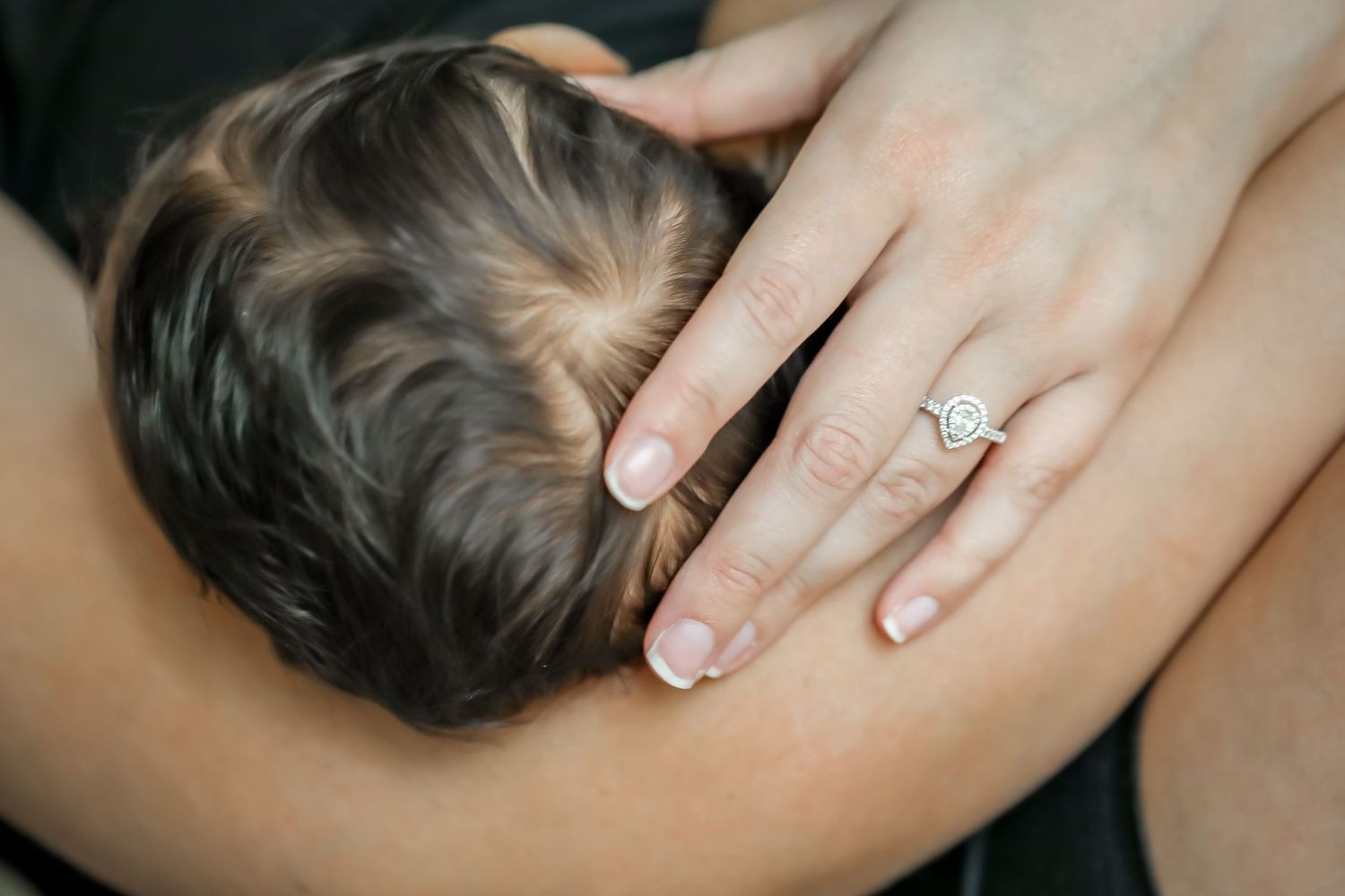 A hand wearing an elegant engagement ring gently holding her baby's head.
