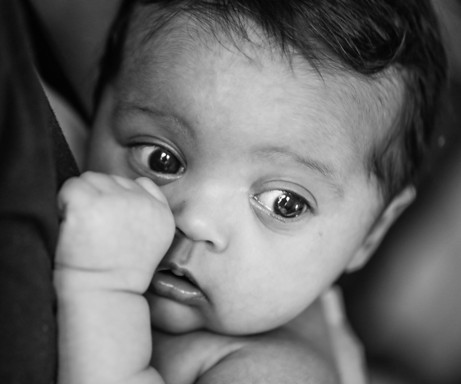 A close-up black and white portrait of a baby with wide eyes resting their hand near their face.