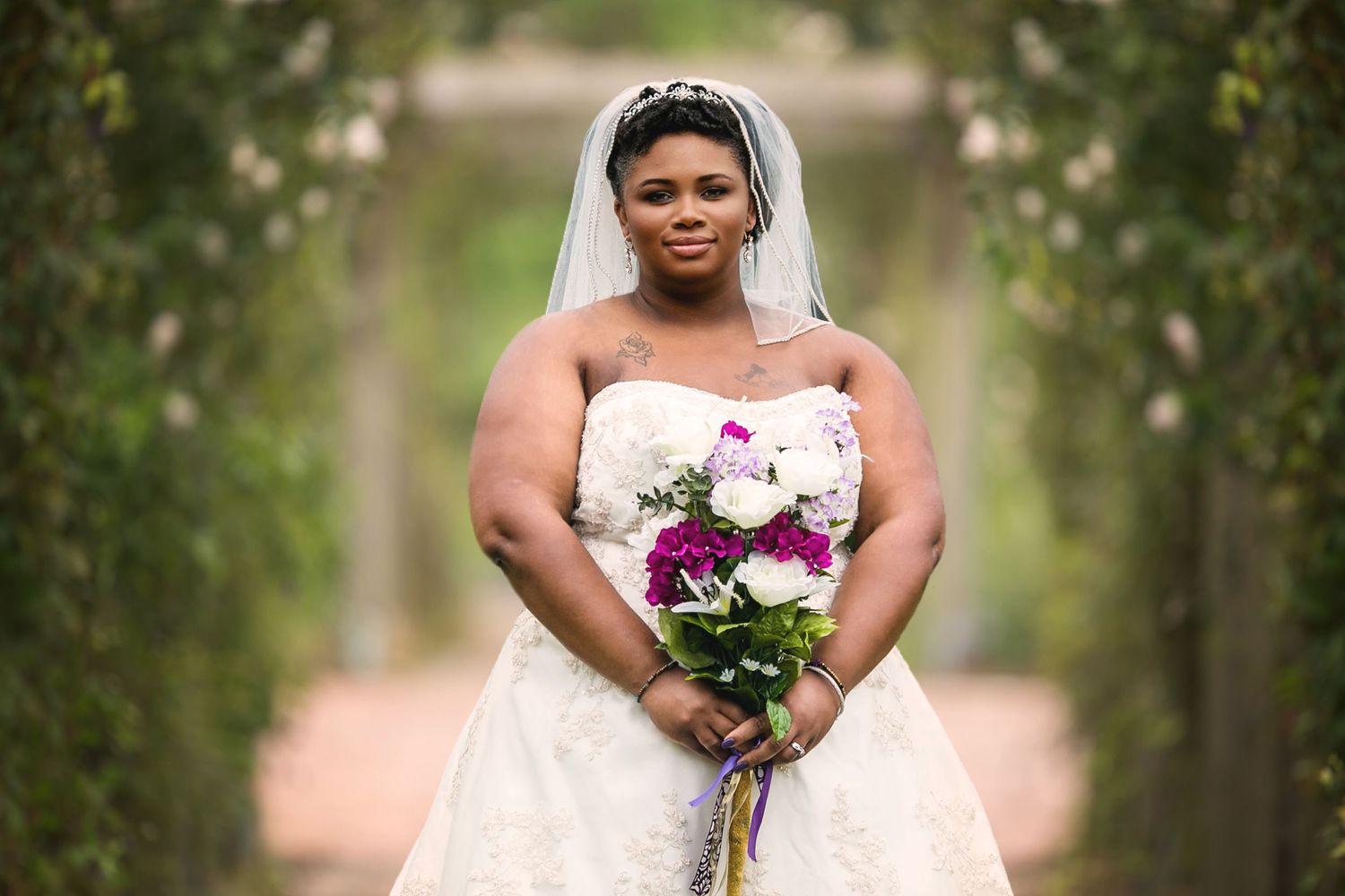 A bride in a strapless white wedding gown holds a purple and white bouquet in garden setting.