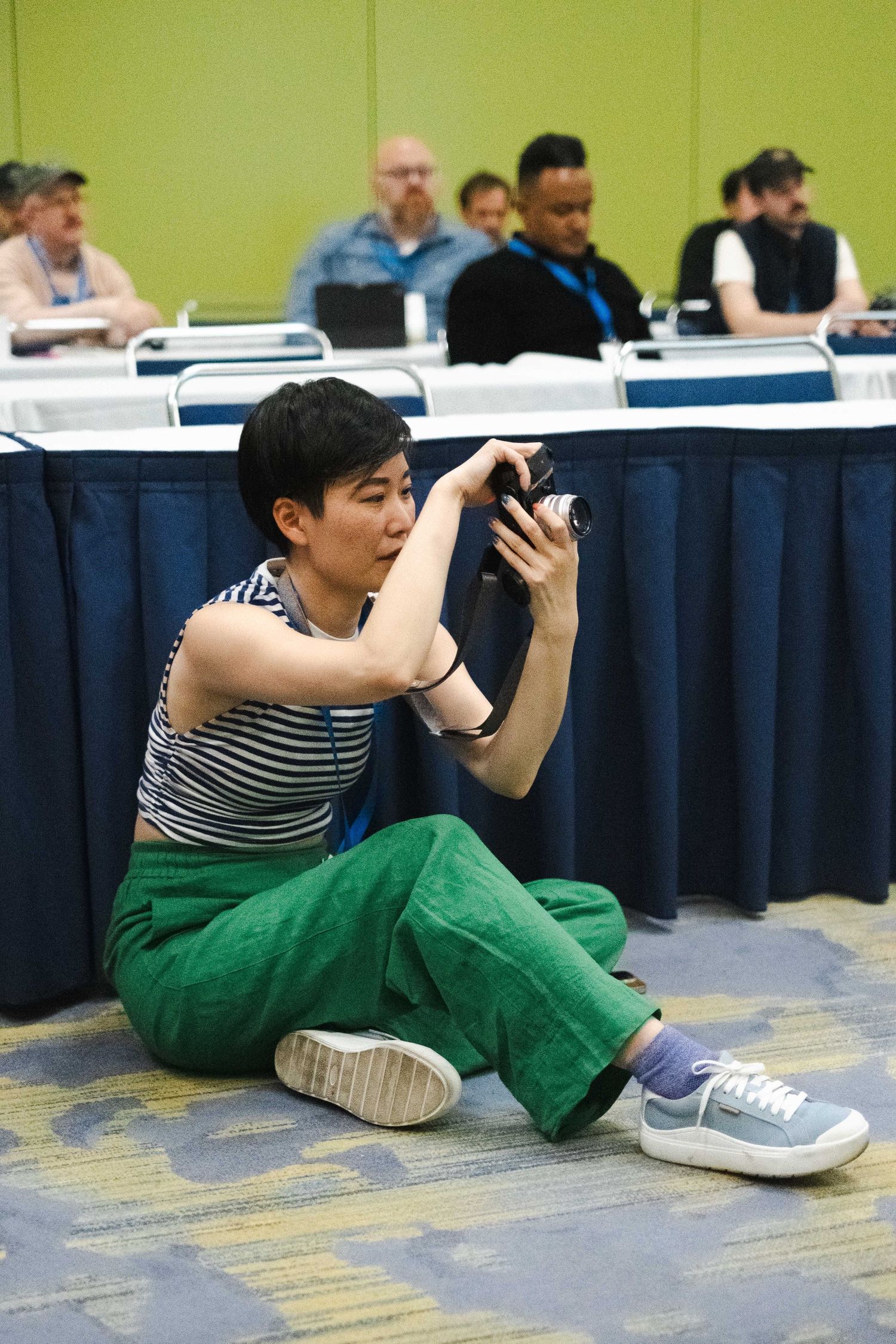 Person in green pants and striped shirt sitting on floor taking photos.