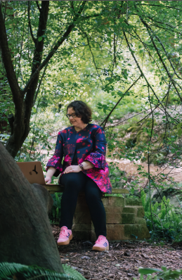 Person wearing colorful floral top sits contemplatively on stone steps surrounded by lush green foliage in park setting.
