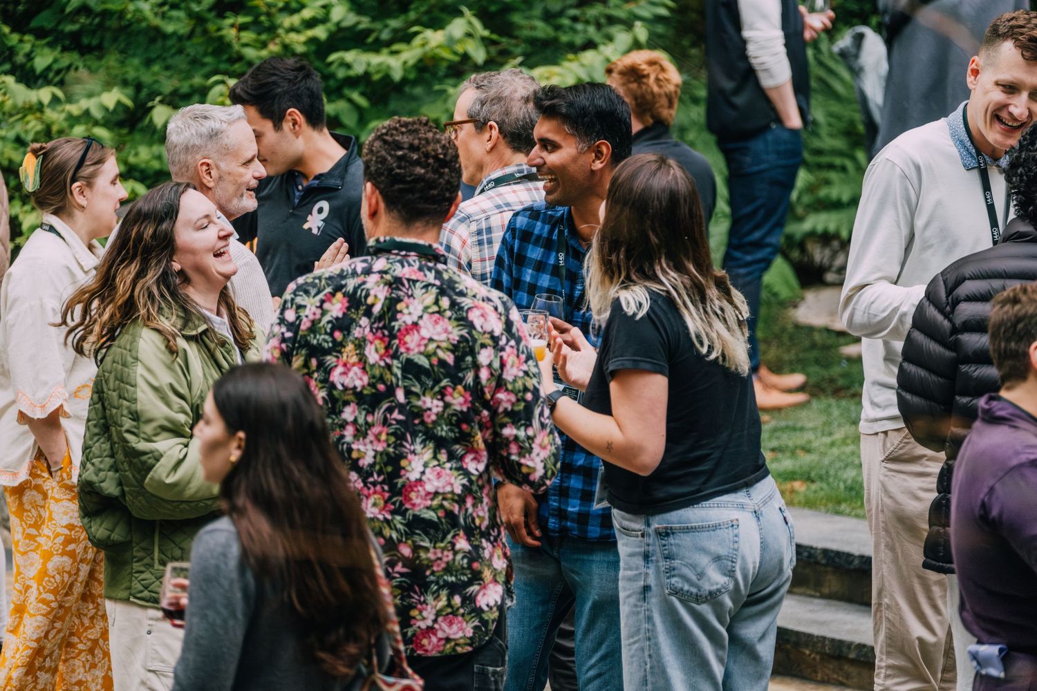 Diverse group of people socializing outdoors at a casual gathering, laughing and conversing in a lush green setting.