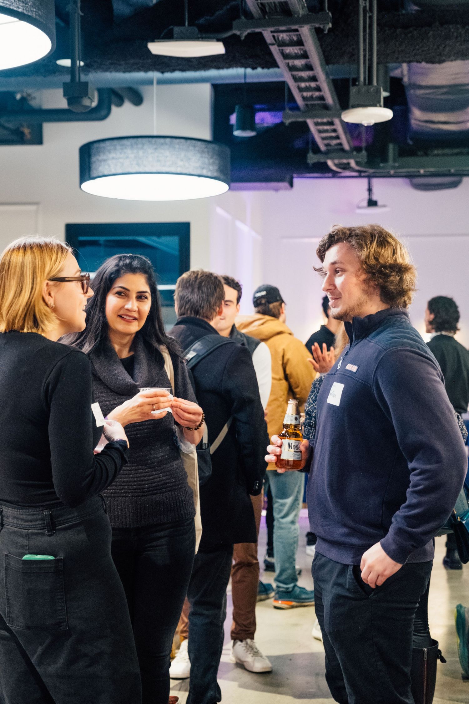 People networking and socializing at an indoor event space with industrial ceiling.