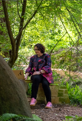 A person sits on a wooden bench surrounded by lush green foliage in a peaceful garden setting.