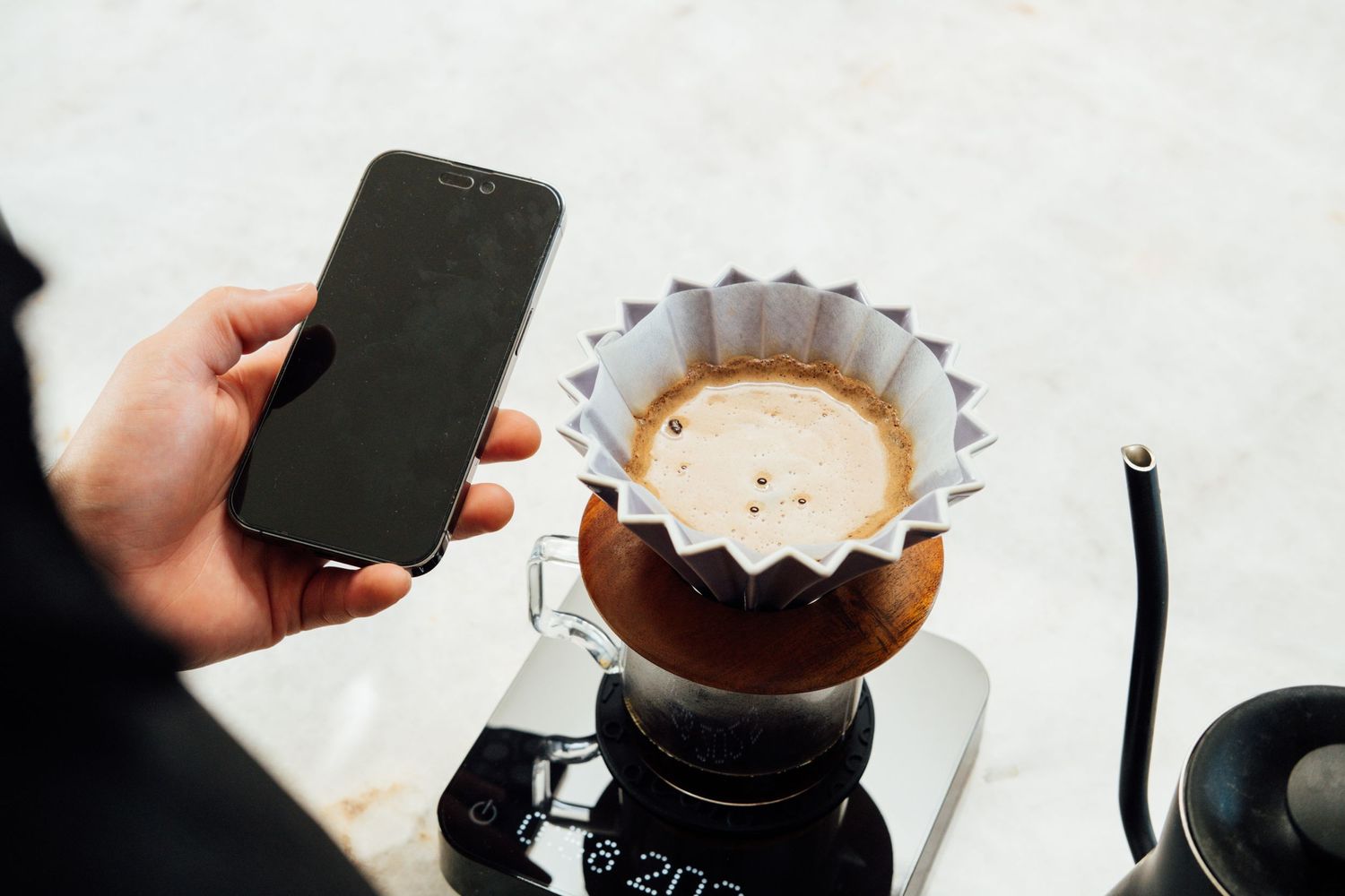 Hand holds smartphone while preparing pour-over coffee with paper filter and black coffee maker.