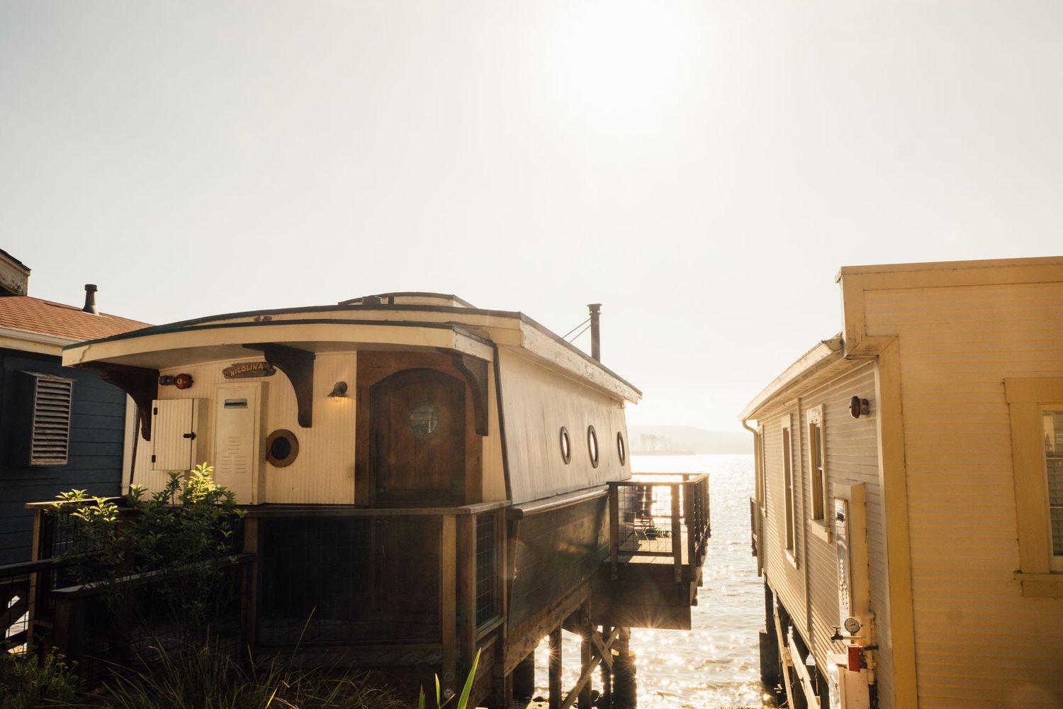 Row of wooden houseboats with sandy brown exteriors and angled roofs silhouetted against a bright overcast sky.