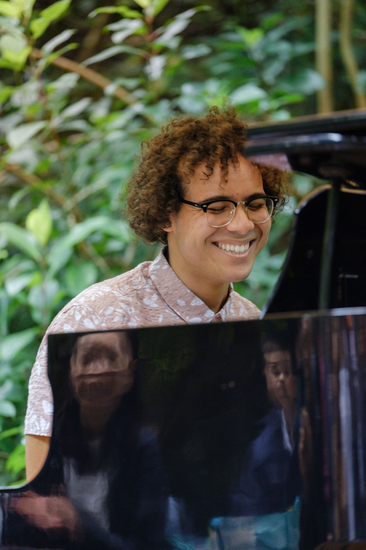 Musician playing piano outdoors with greenery in the background.