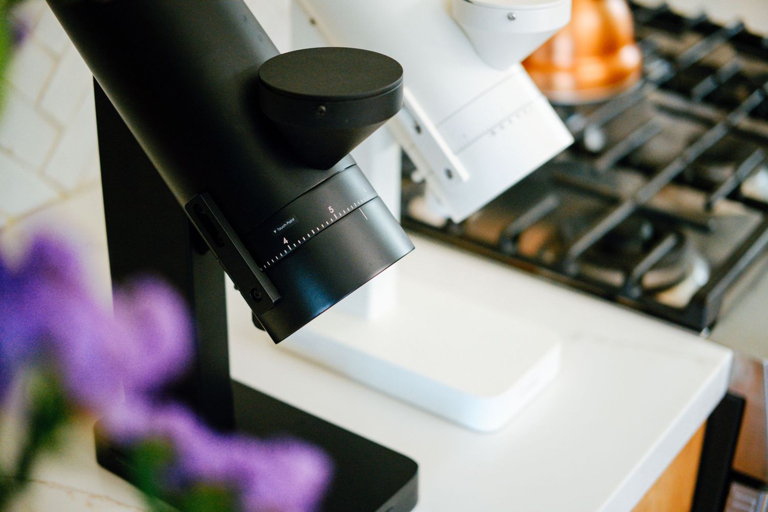 Close up of black coffee grinder next to stove with purple flowers in foreground.
