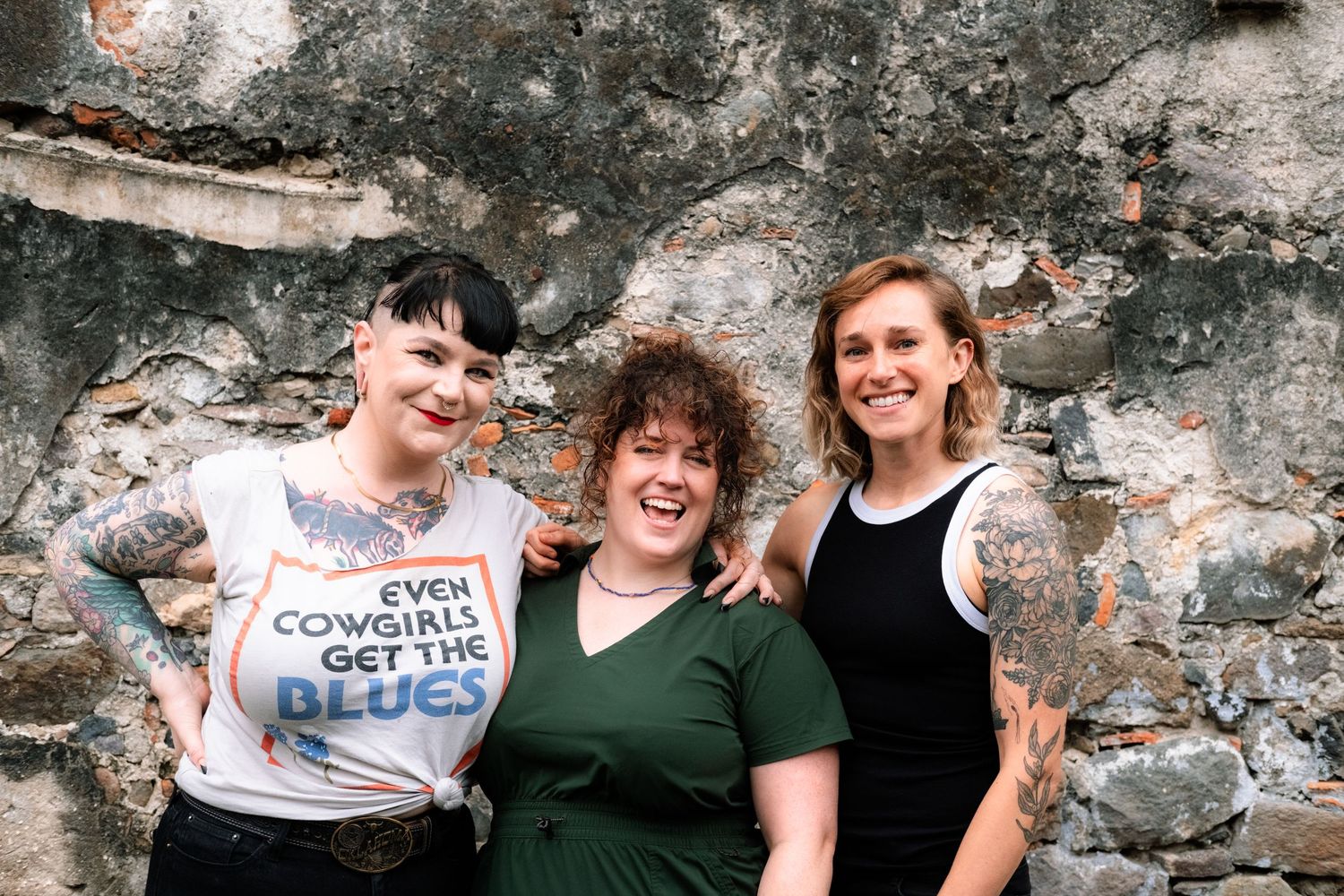 Three friends posing together against a weathered stone wall background in casual attire.