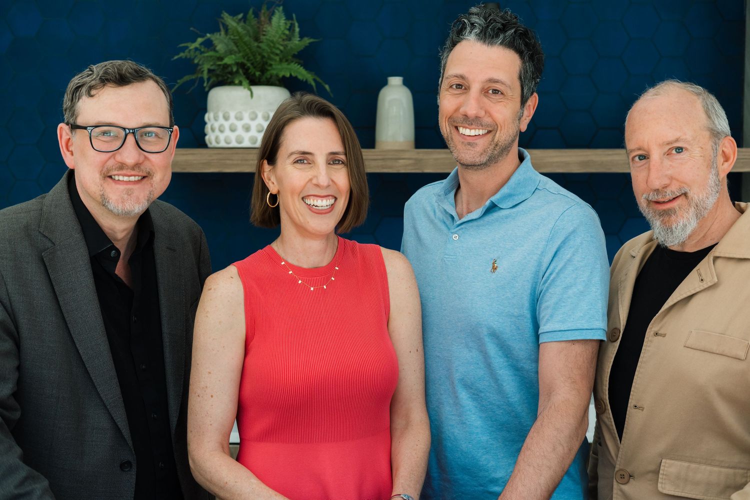 Four people stand together in front of a blue wall, with one wearing a red dress and others in casual and business attire.