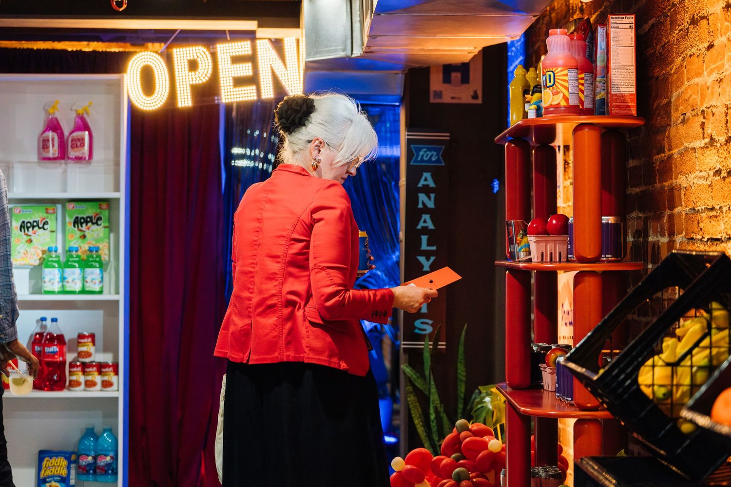 Elderly woman in red jacket browsing colorful shelves in a neon-lit convenience store with an OPEN sign.