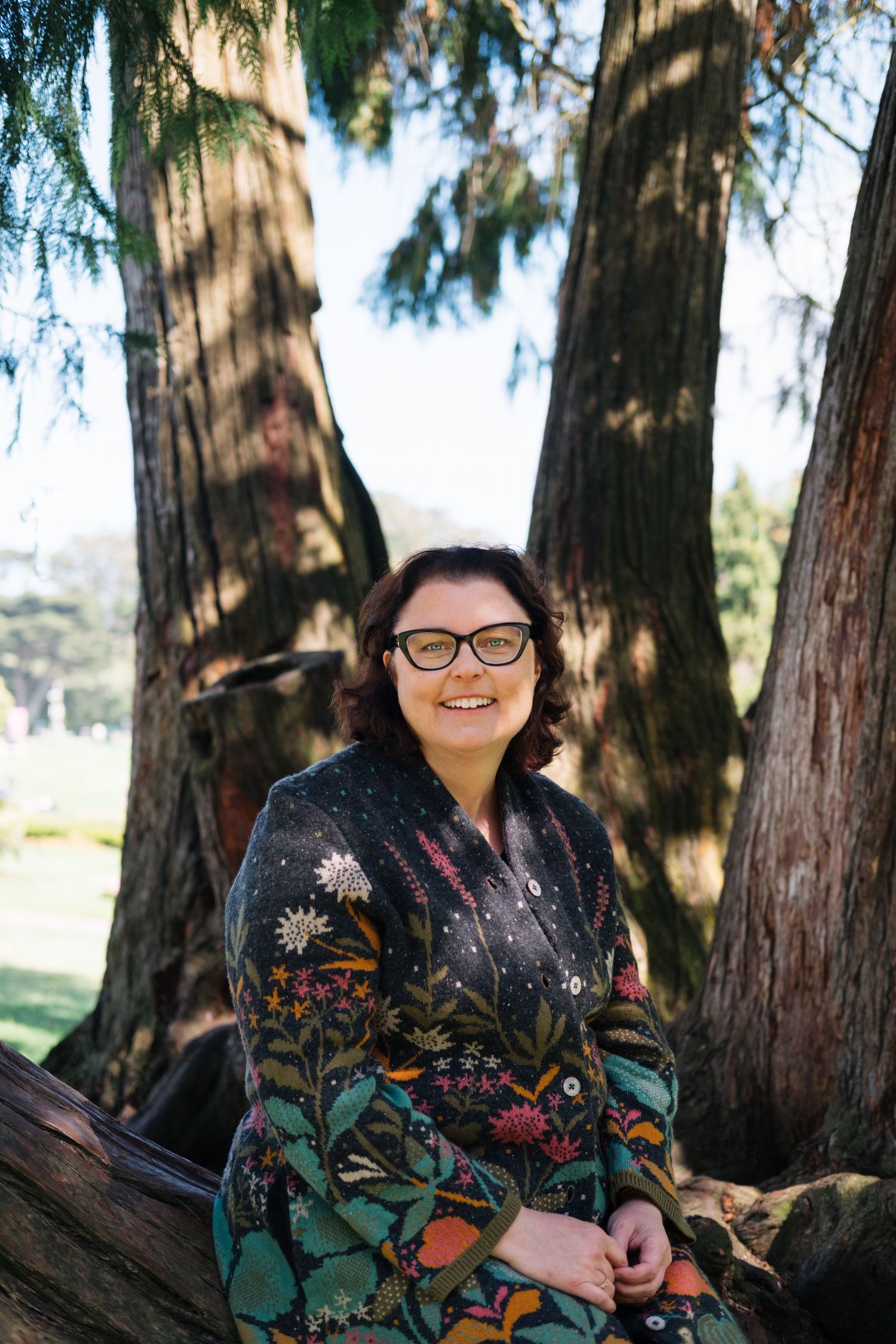 A person wearing a floral patterned dress sits among tall cypress trees in a park setting.