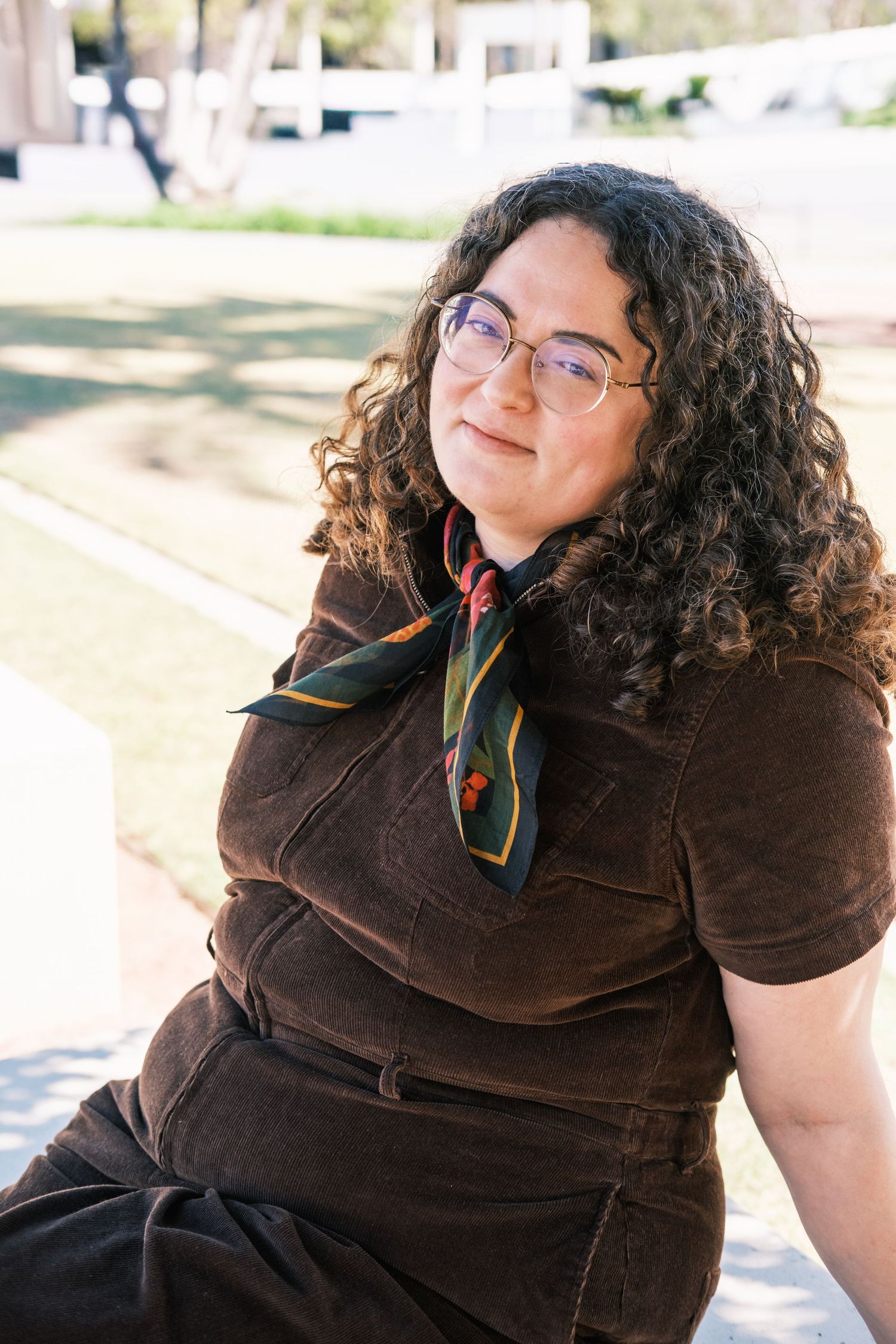 Young woman with curly hair and glasses wearing a brown corduroy outfit and colorful scarf outdoors.