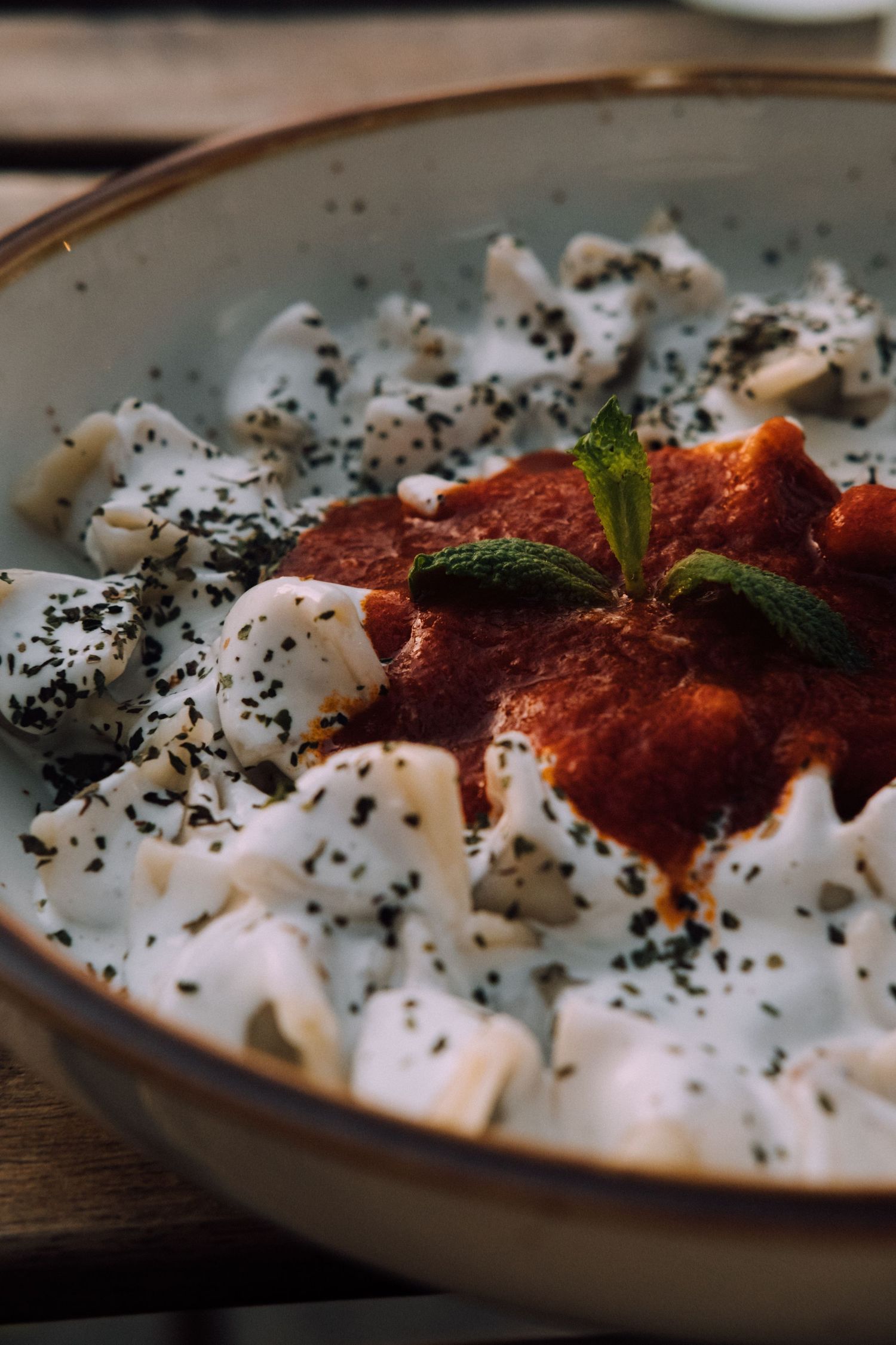 A bowl of creamy sauce topped with red tomato sauce, black sesame seeds and fresh herbs against a wooden surface.