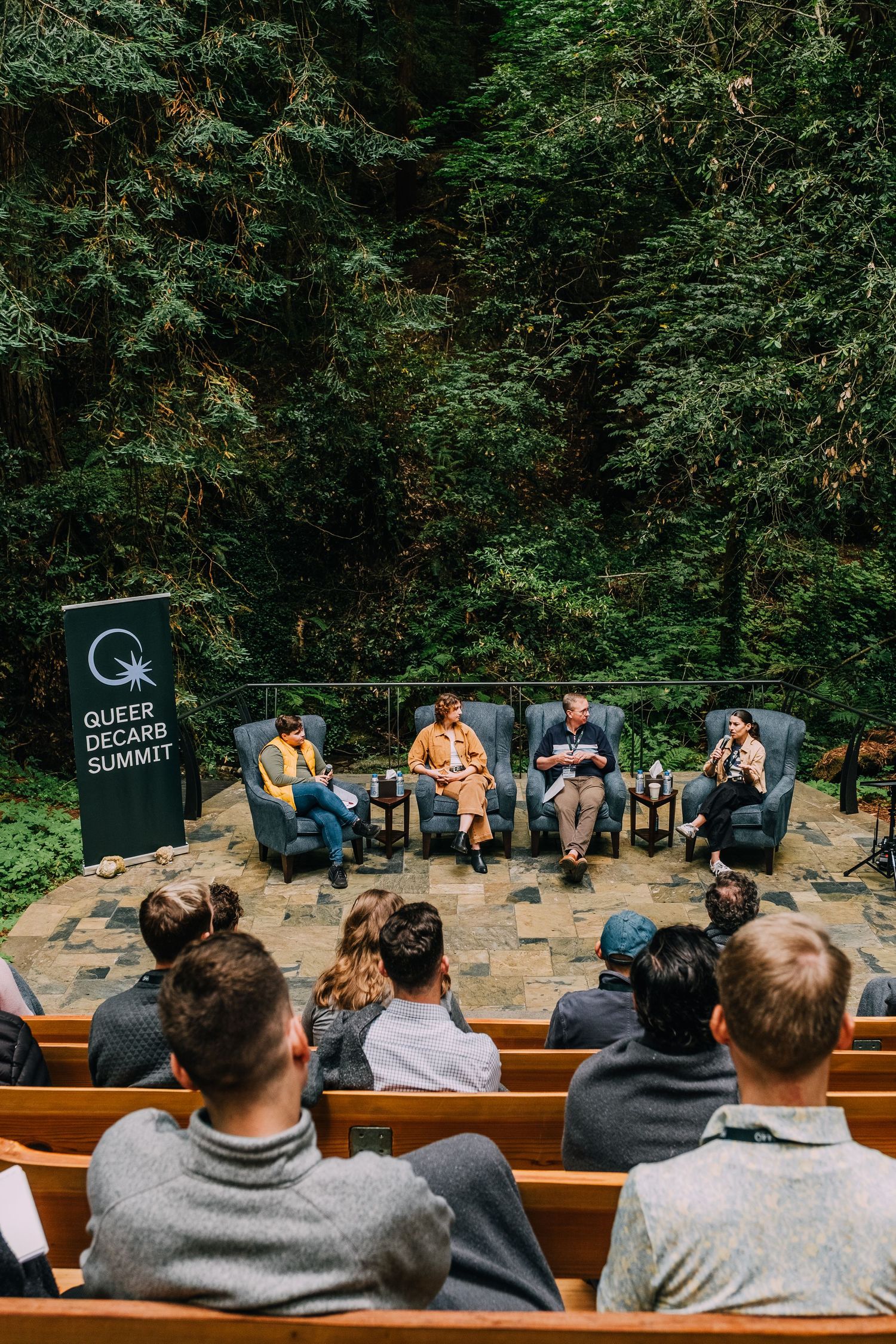 Panelists speak at the Queer Decarb Summit outdoor amphitheater surrounded by lush green forest trees.