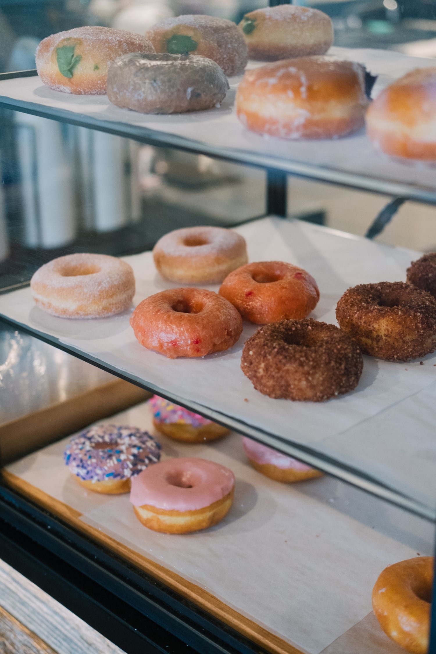 Assorted fresh donuts with different toppings and glazes displayed in a bakery case on parchment paper.