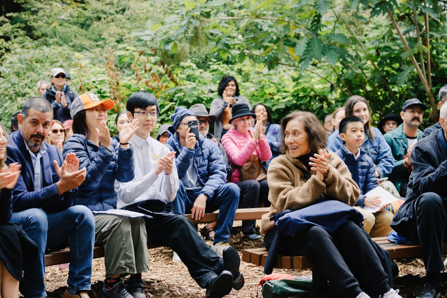 Audience seated outdoors on wooden benches in a natural setting during an event.