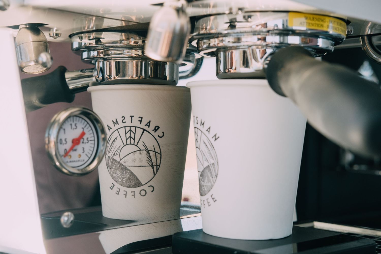 Close-up of coffee machine pressure gauges and branded cups in a cafe setting.