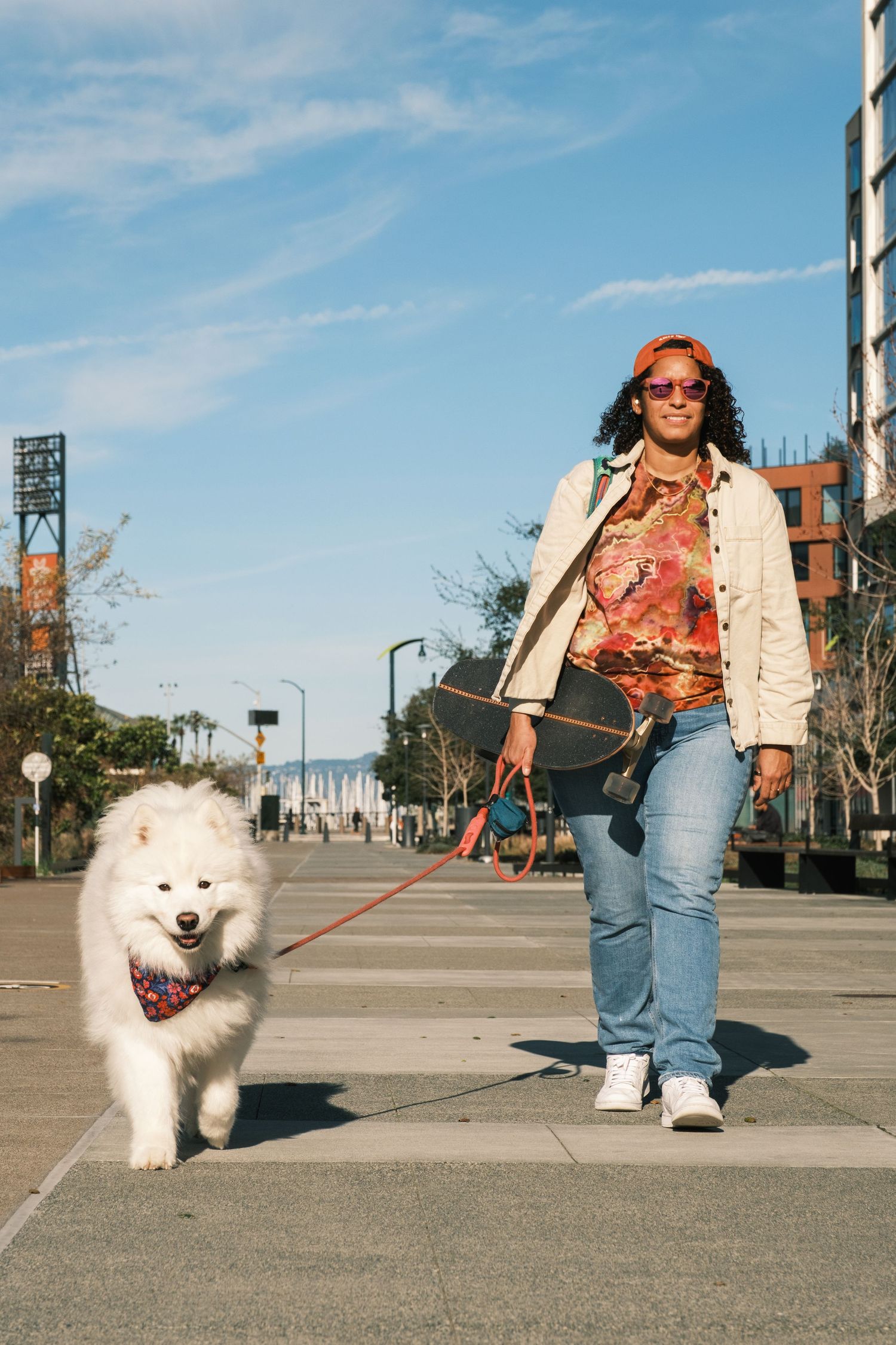 Someone walking a fluffy white Samoyed dog on a city sidewalk near a baseball stadium on a sunny day.