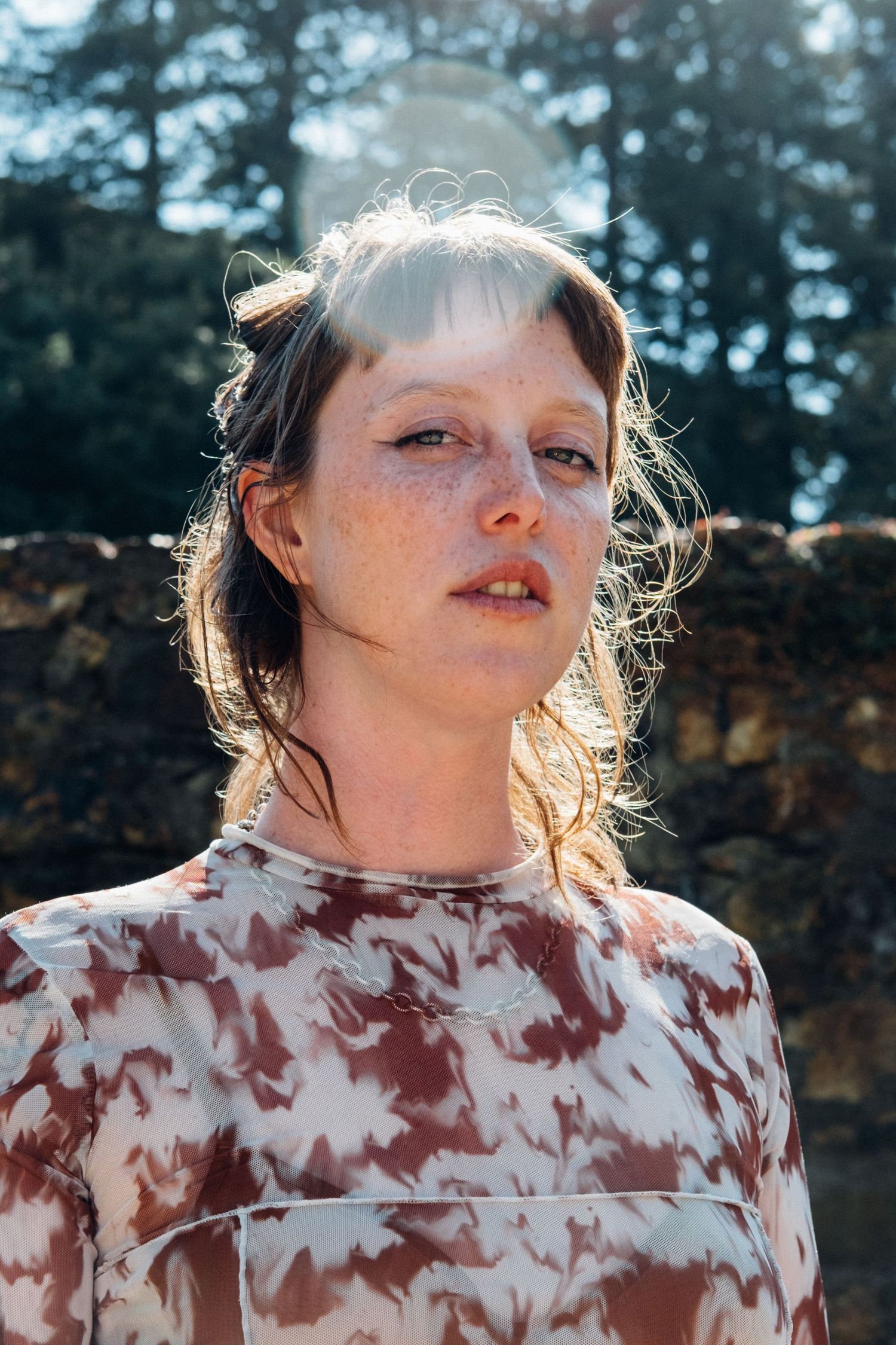 A person wearing a white and burgundy tie-dye shirt poses against a natural outdoor backdrop in warm sunlight.