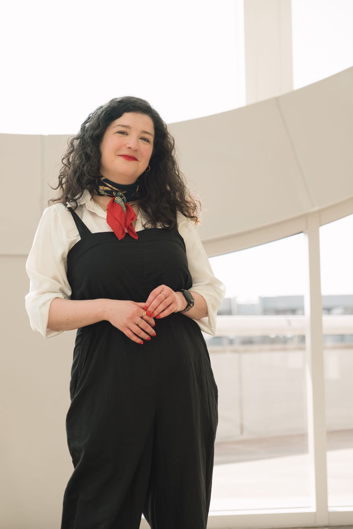 Woman in black overalls, white blouse, and red neckerchief posing indoors with curly dark hair and red lipstick.