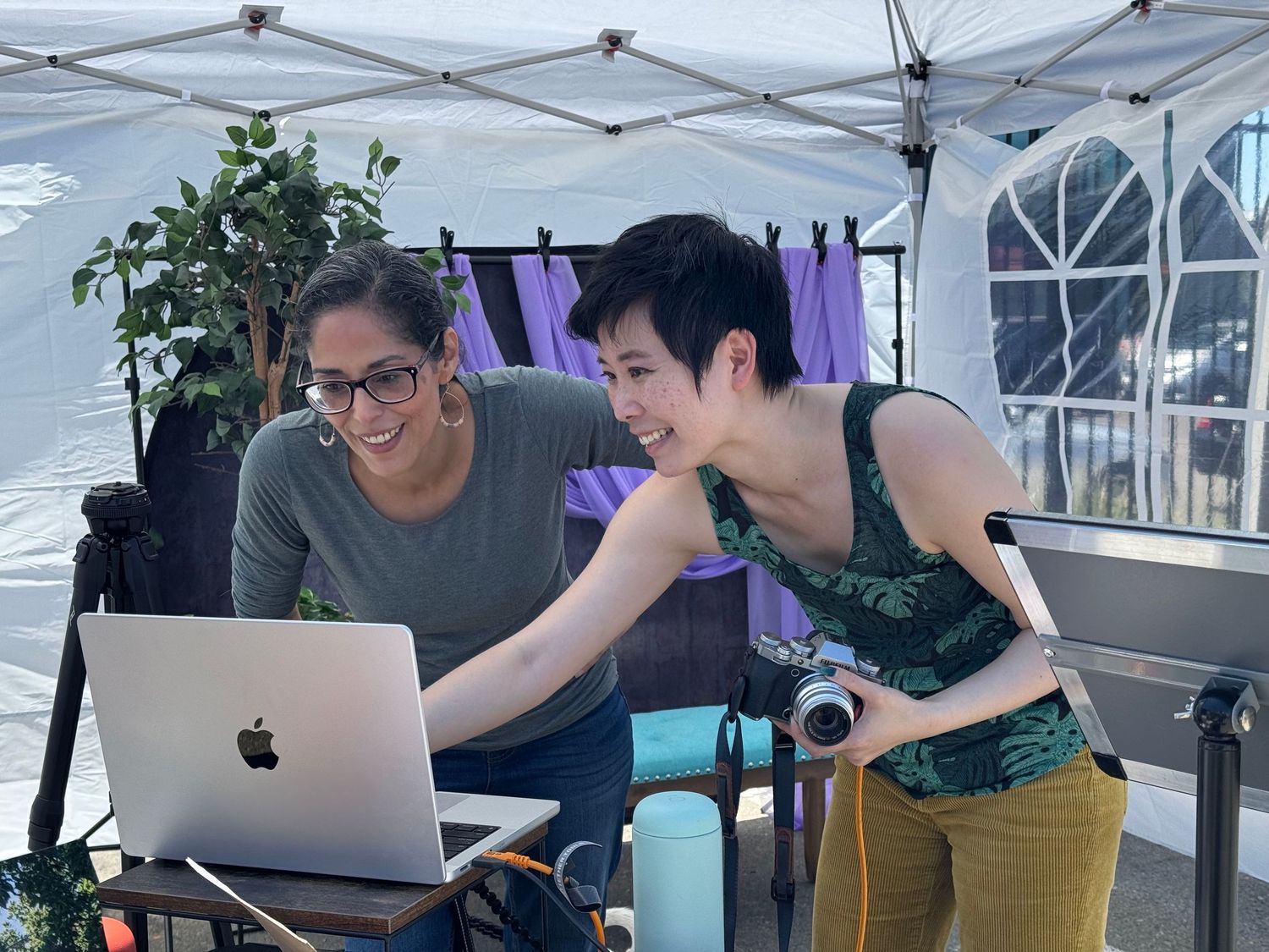 Two women smiling and collaborating on a laptop under a white tent outdoors.
