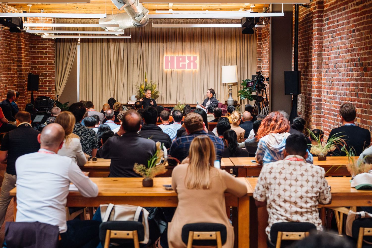 Audience seated at wooden tables facing speakers at a DEX event in a brick-walled venue with warm lighting.