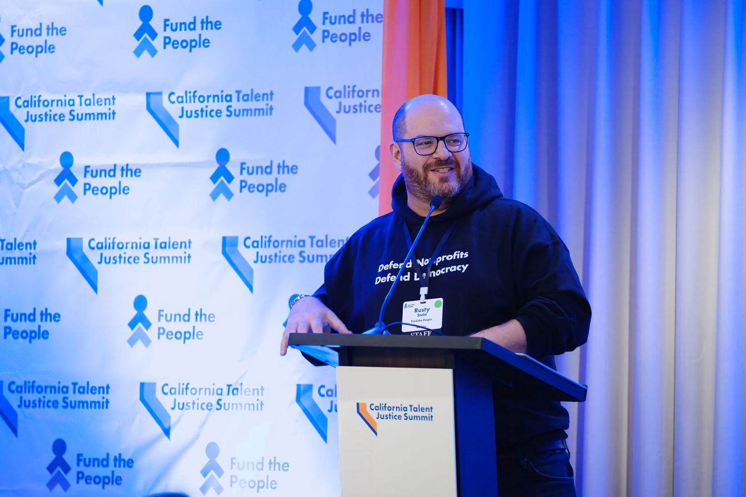 Speaker presenting at conference podium with branded blue backdrop displaying organizational logos.