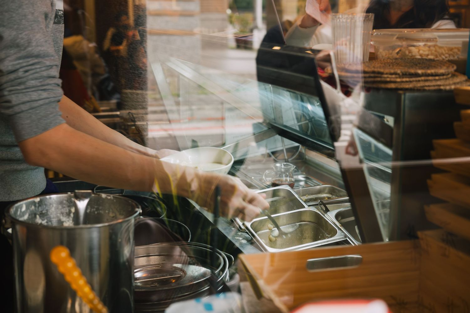 Barista preparing drinks at a coffee shop counter with equipment and supplies visible through glass display case.