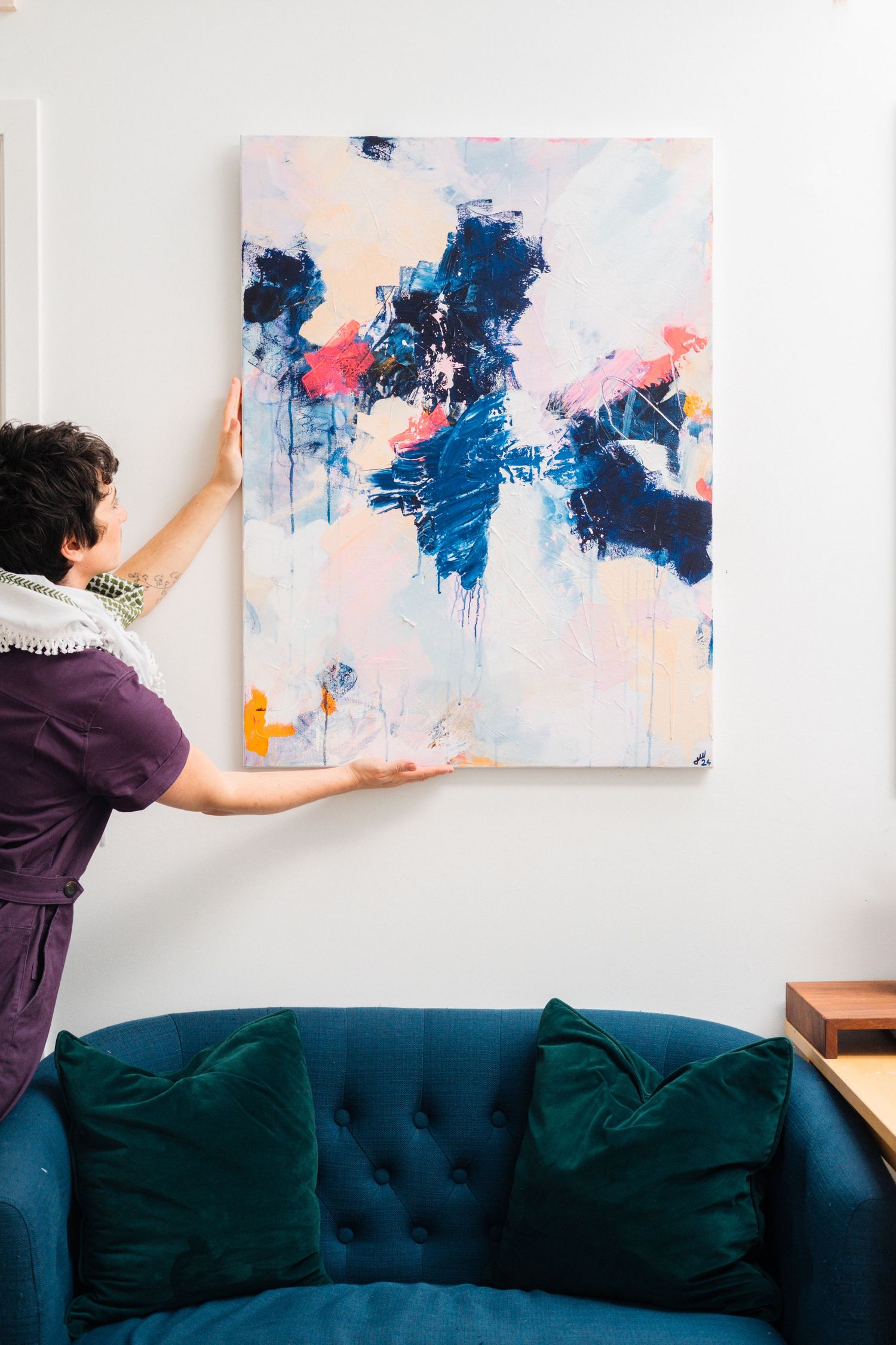Abstract blue and pink artwork being hung on wall above dark teal couch.