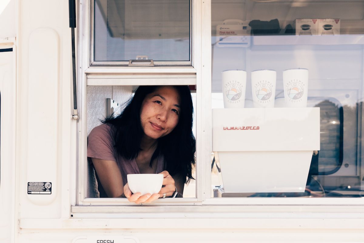 Someone smiles while working in a white service window counter with takeout containers stacked nearby.