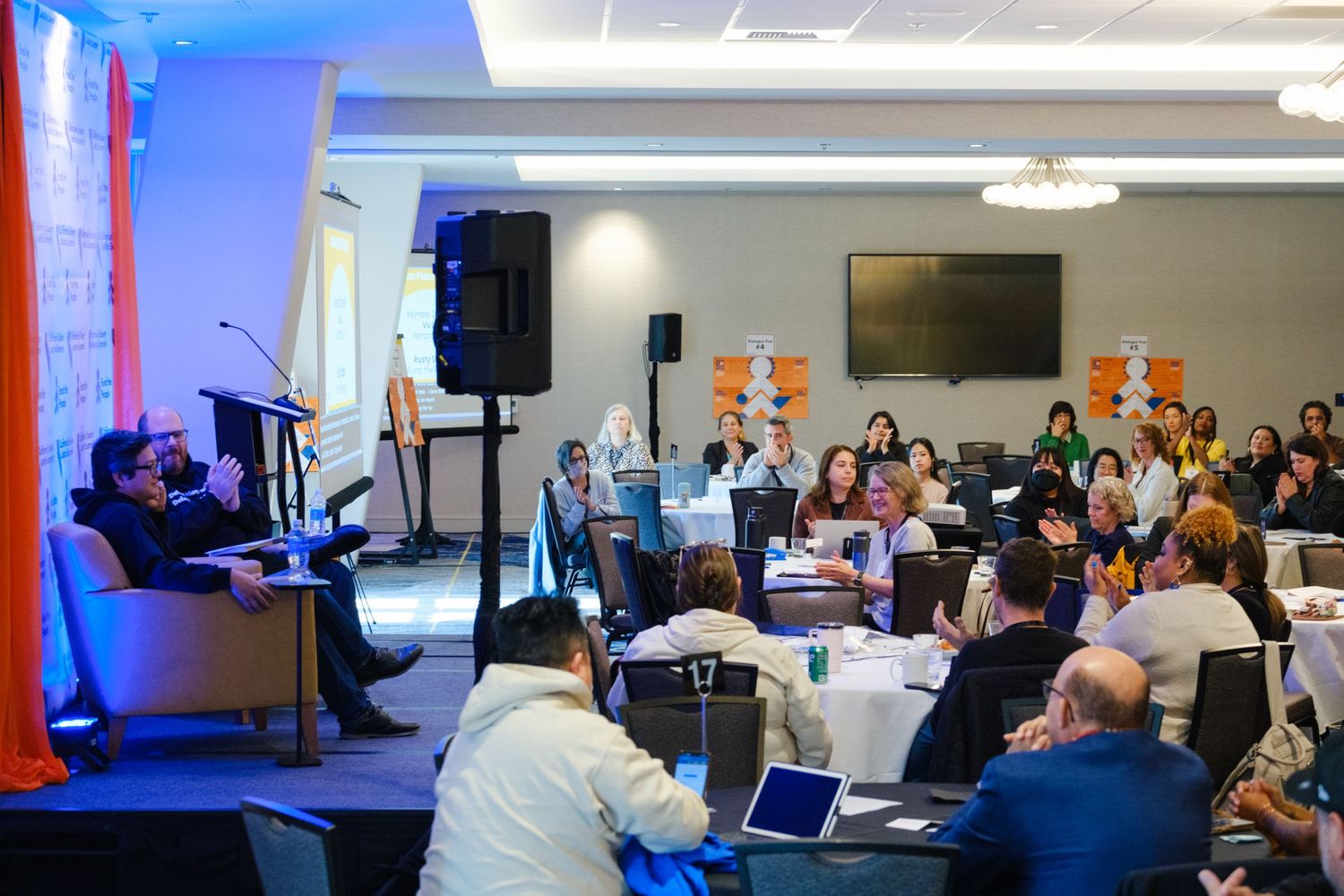 Wide view of a conference room with audience members seated at round tables during a presentation.