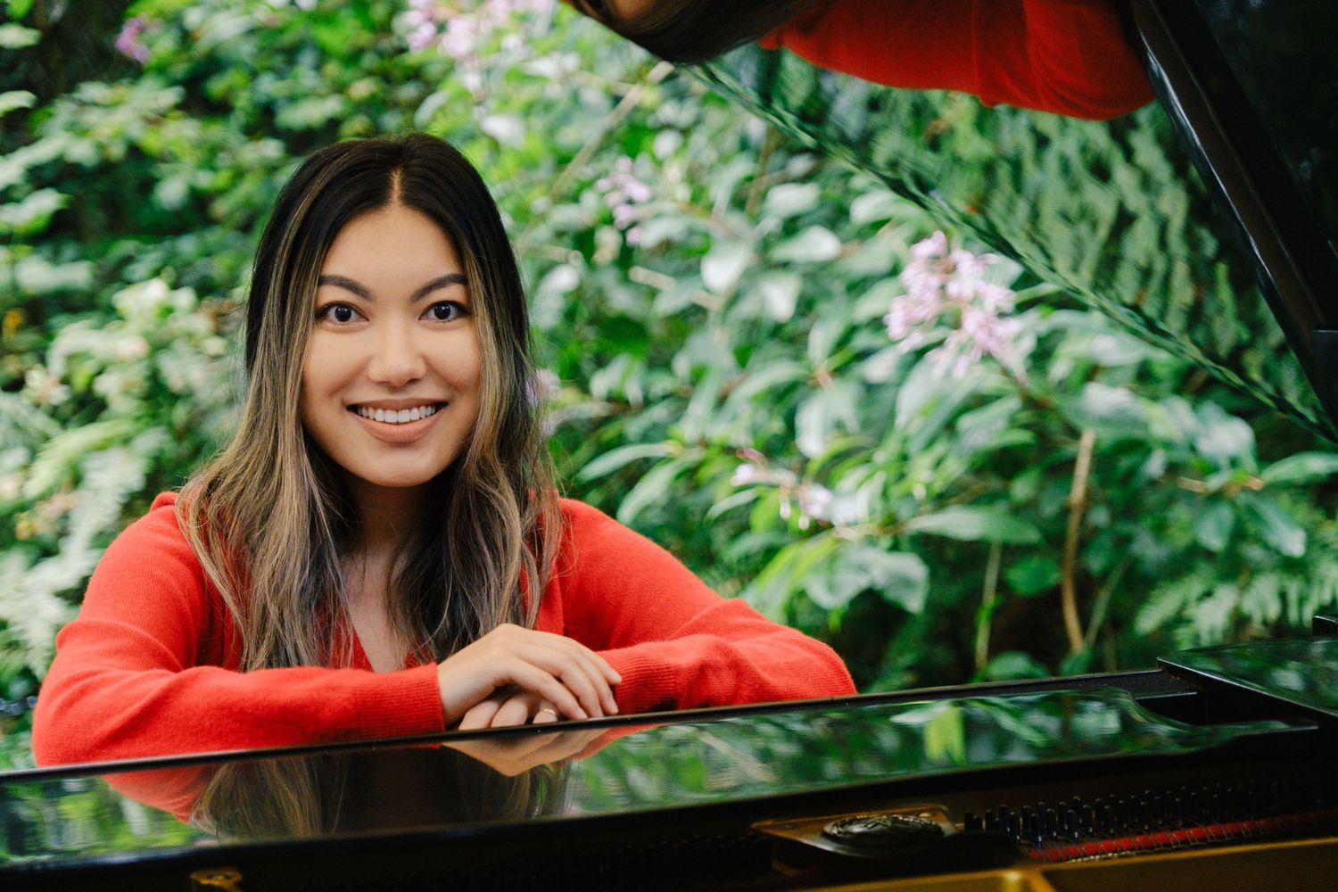 Woman in bright red sweater plays piano outdoors surrounded by lush green foliage and natural light.