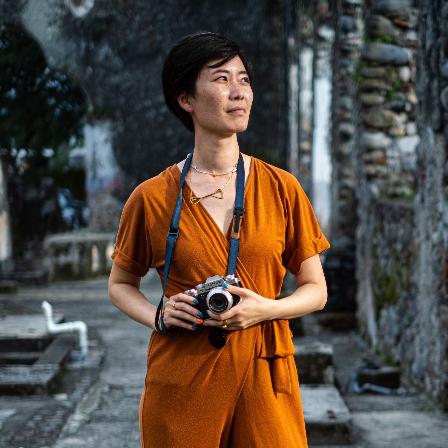 A woman in an orange jumpsuit holds a camera outdoors near stone ruins, looking thoughtfully into the distance.