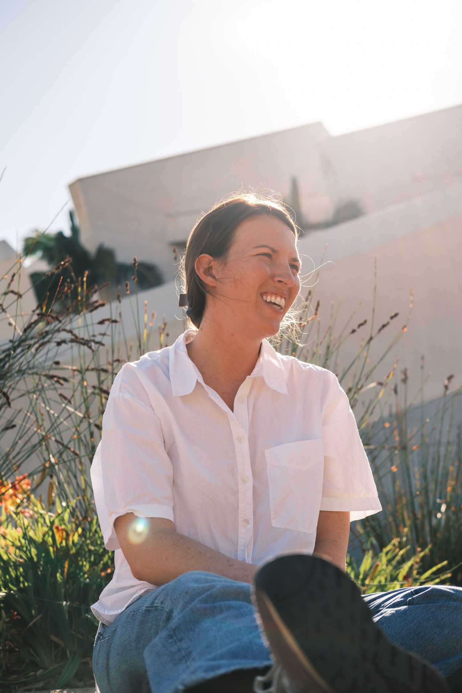 Happy woman in white linen shirt sitting outdoors in bright sunlight near a white wall.