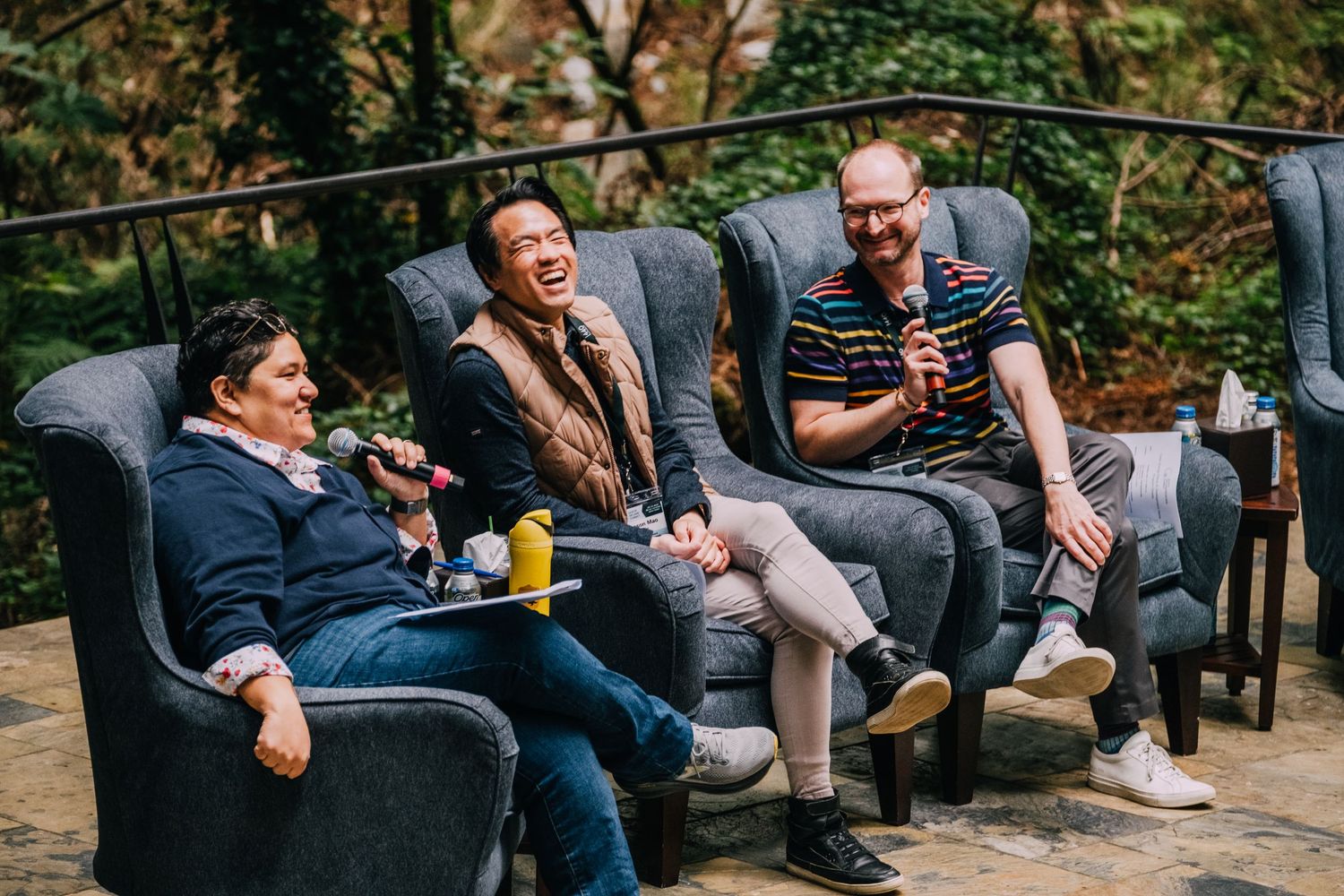 Three men laughing and relaxing in gray armchairs outdoors at a casual panel discussion event.