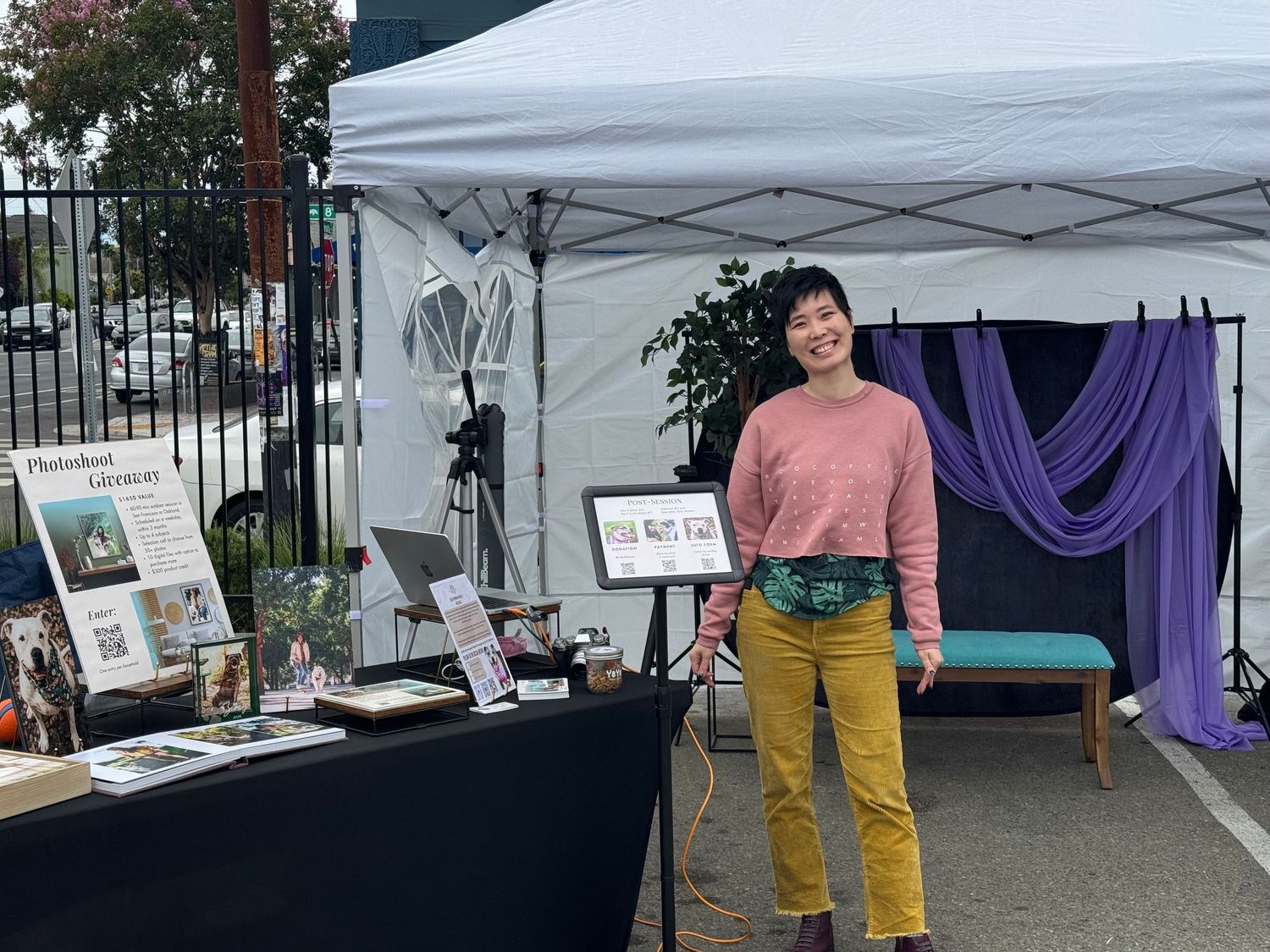 Person standing at outdoor market booth with books and art displays under white canopy tent with purple drape backdrop.