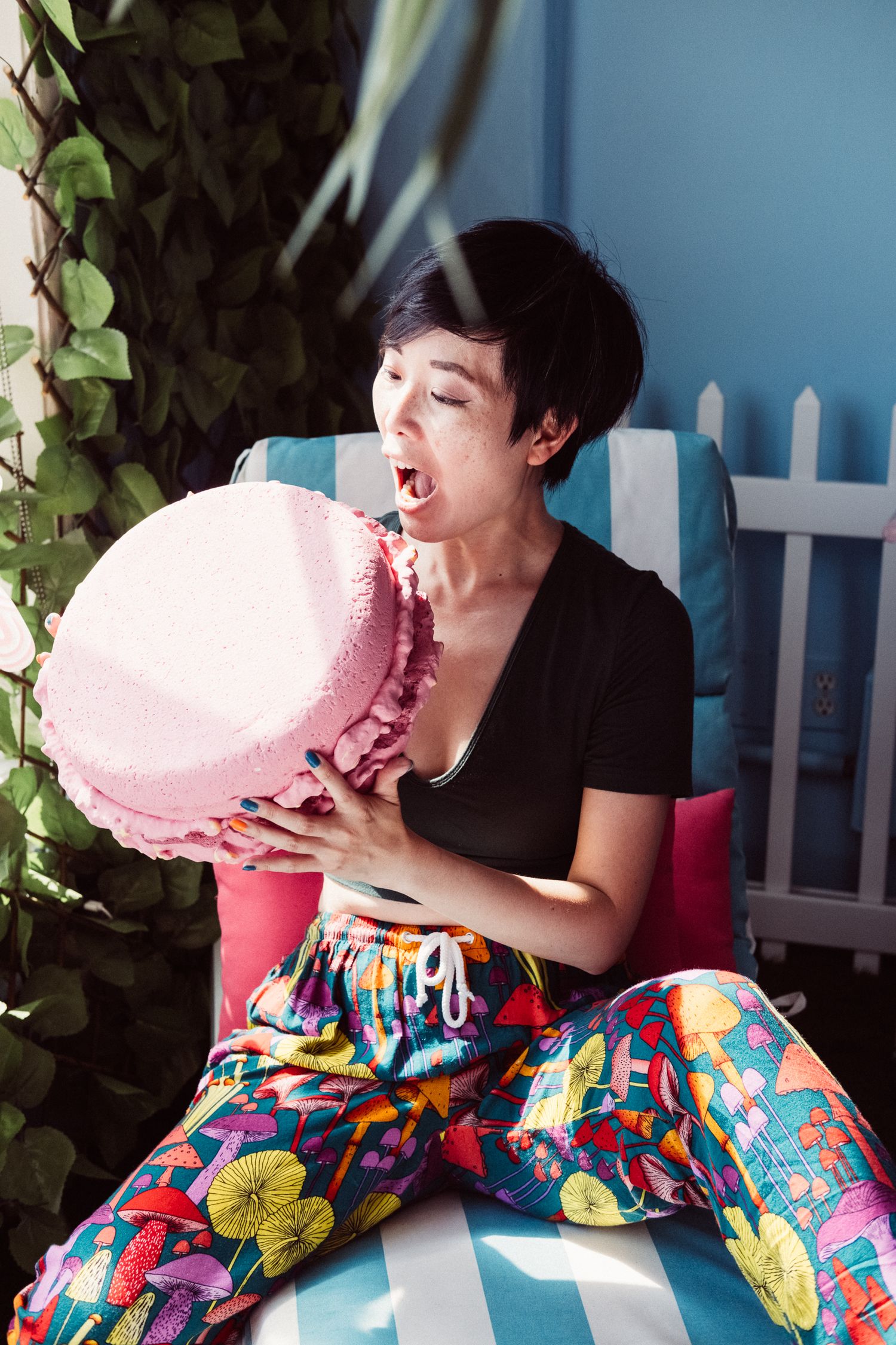 Someone wearing colorful patterned pants bites into a large pink cotton candy while sitting outside on a sunny day.