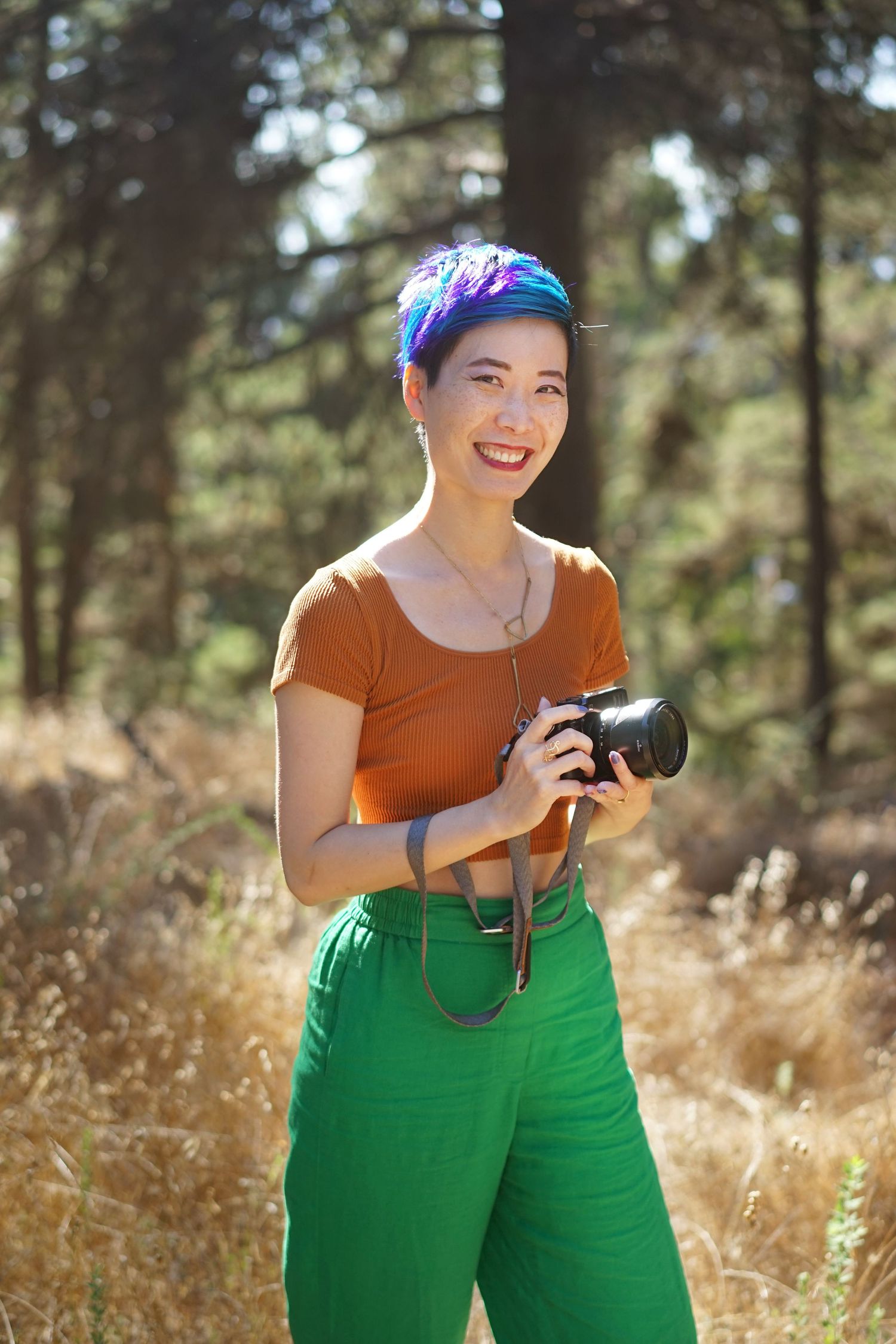 Person in orange top and green pants holding camera in sunny field.