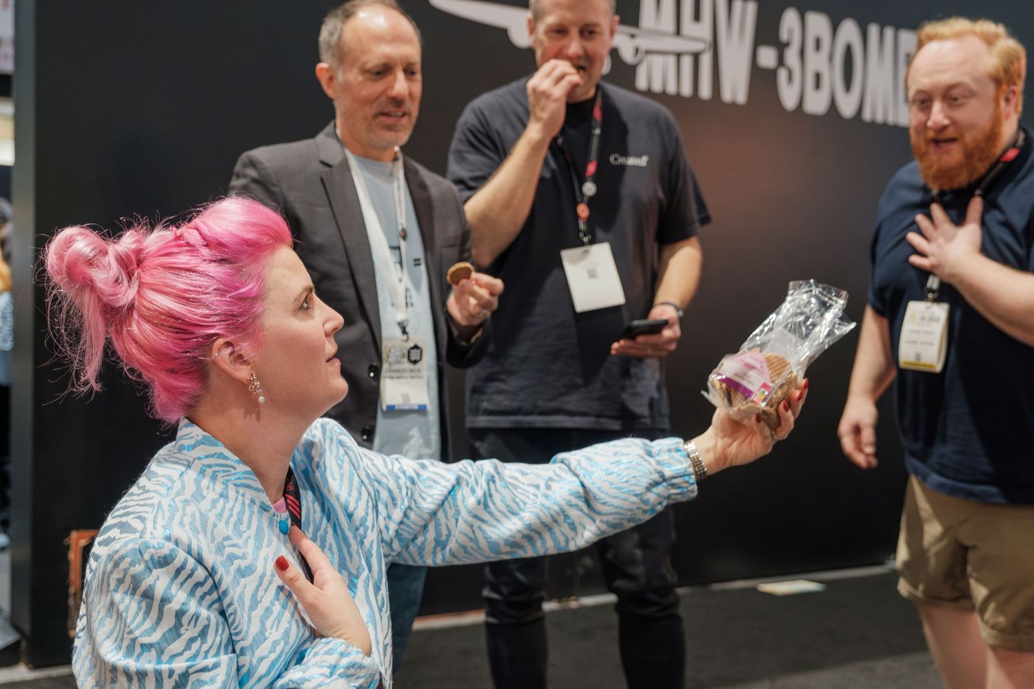 Woman with pink hair holds a product at a trade show booth while onlookers react with surprise.