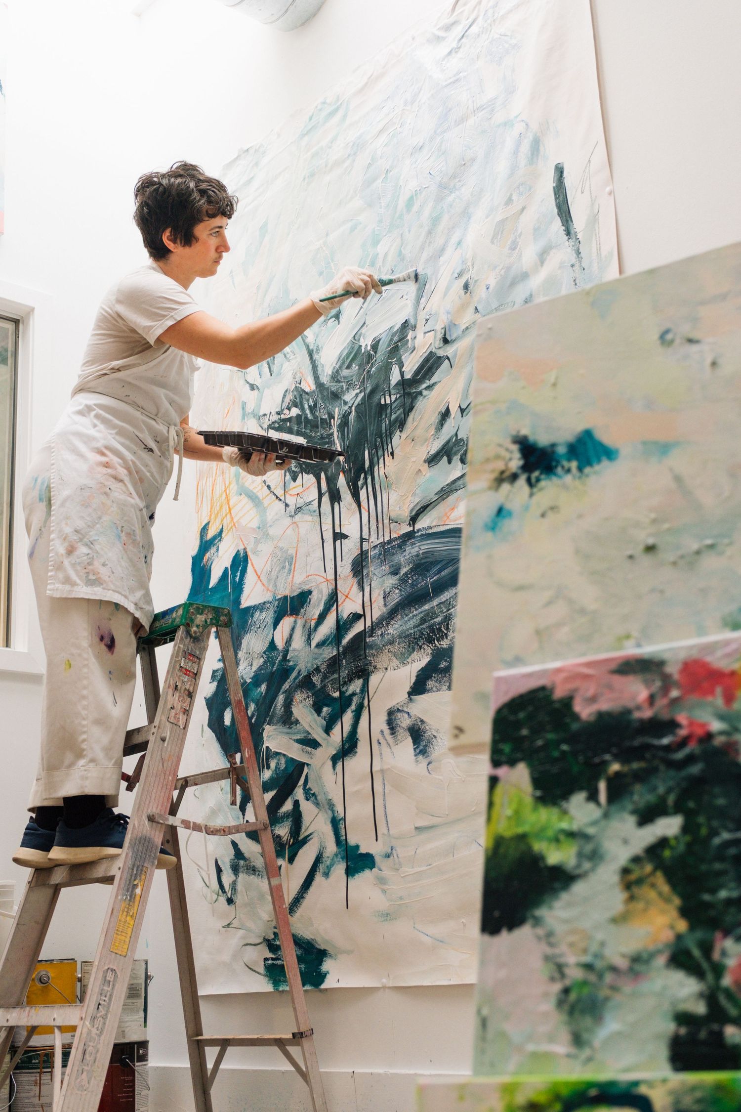 Artist in white outfit works on large abstract painting from ladder in bright studio interior series.