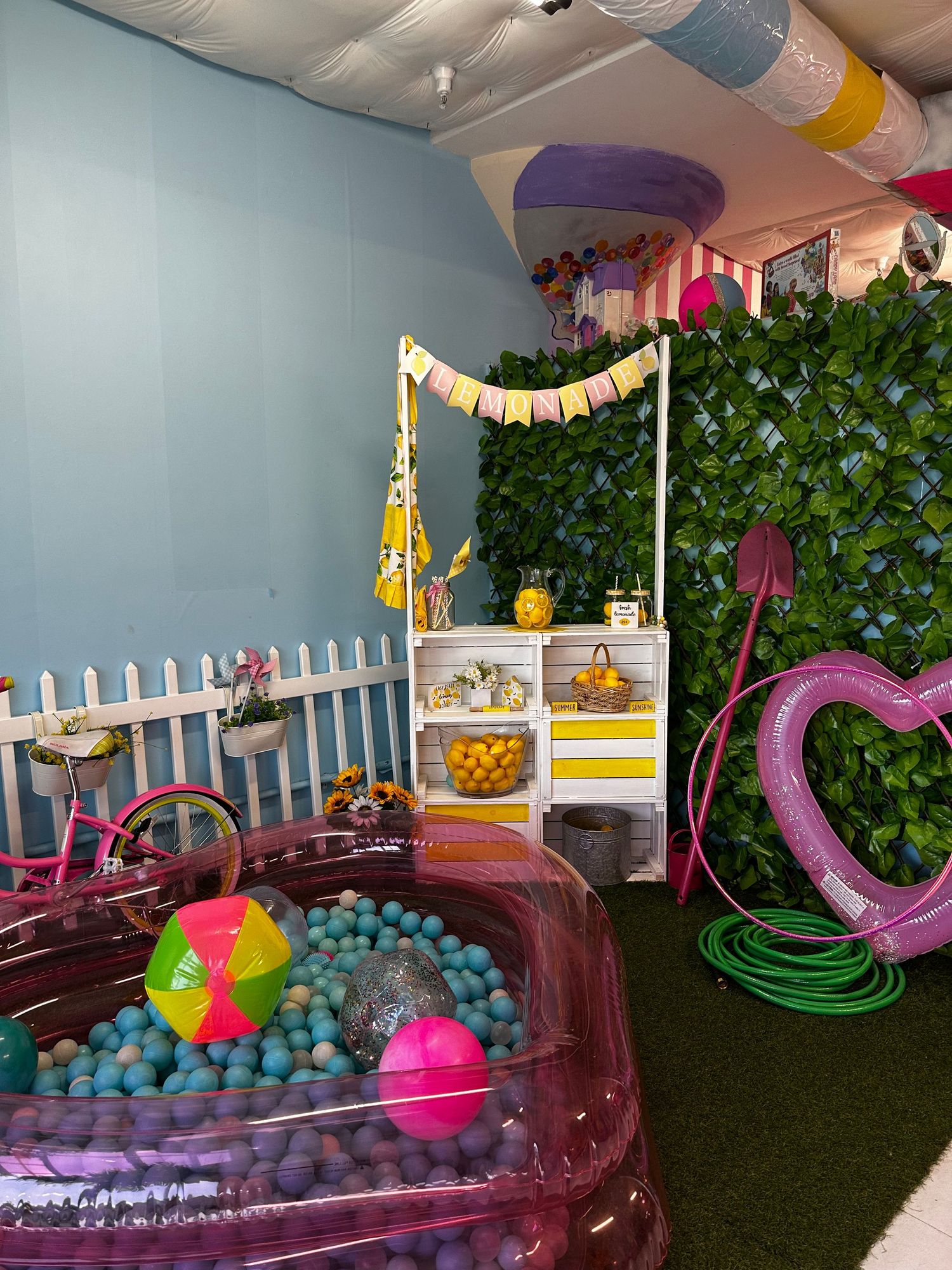 Colorful indoor play area with ball pit, pink decorations, ivy wall backdrop and white picket fence details.